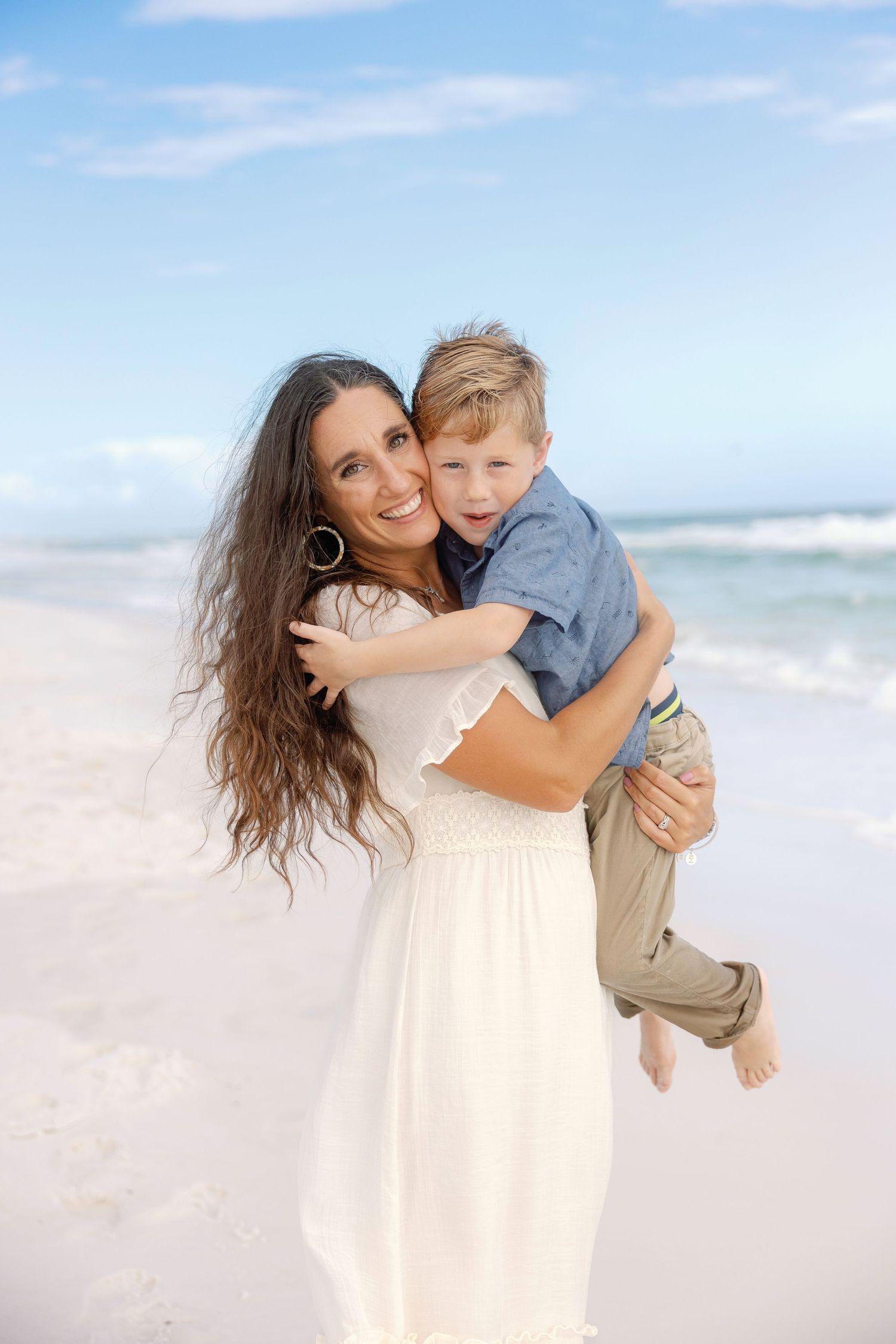 A tender moment shared between parent and child embracing on a sunny beach with ocean waves in the background.