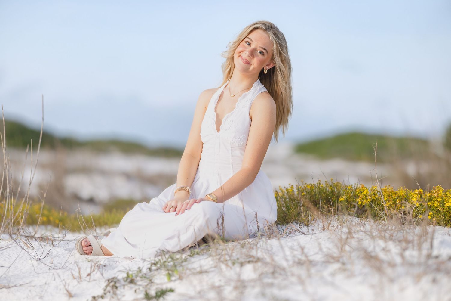 Woman in white dress sitting on white sand dunes with yellow wildflowers on a sunny beach day.