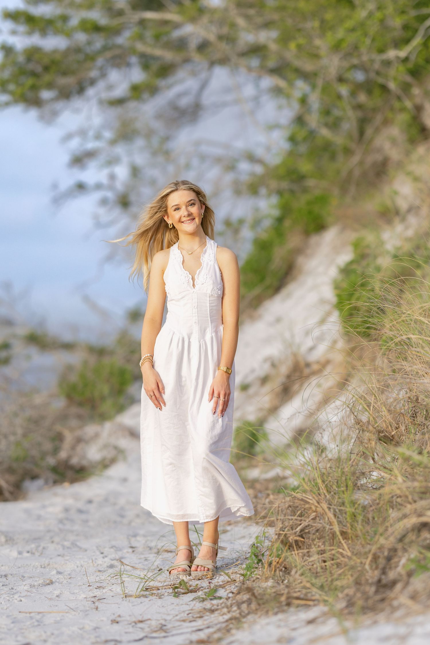 Young blonde woman in white midi dress walks sandy coastal path, smiling with wind-blown hair and green dunes behind her.