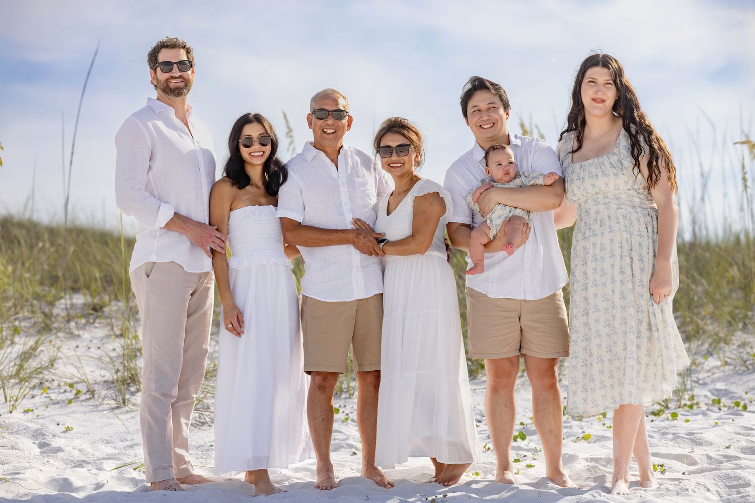 Family of seven posing together on a sandy beach, dressed in white and neutral tones, smiling for a portrait.