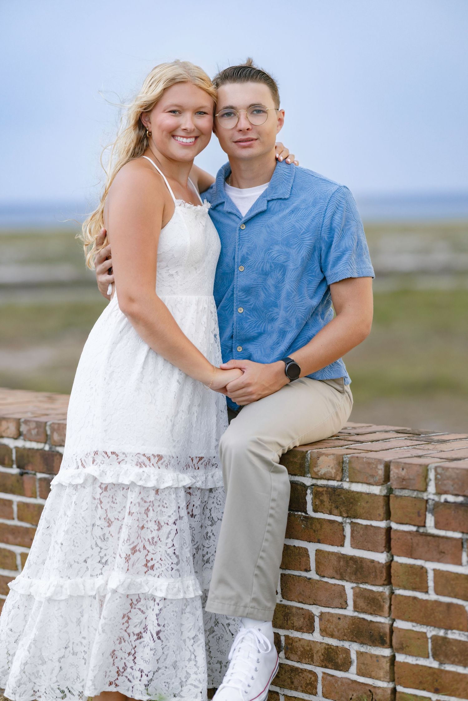 A young couple poses together on a brick wall outdoors, woman in white lace dress and man in blue shirt.