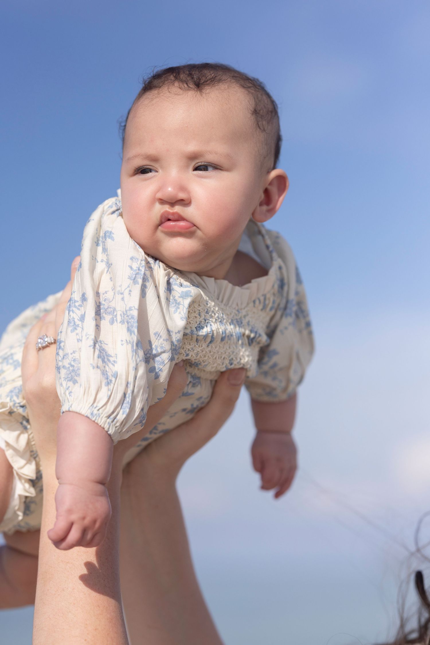 Baby in floral romper lifted in the air against a bright blue sky by mother.
