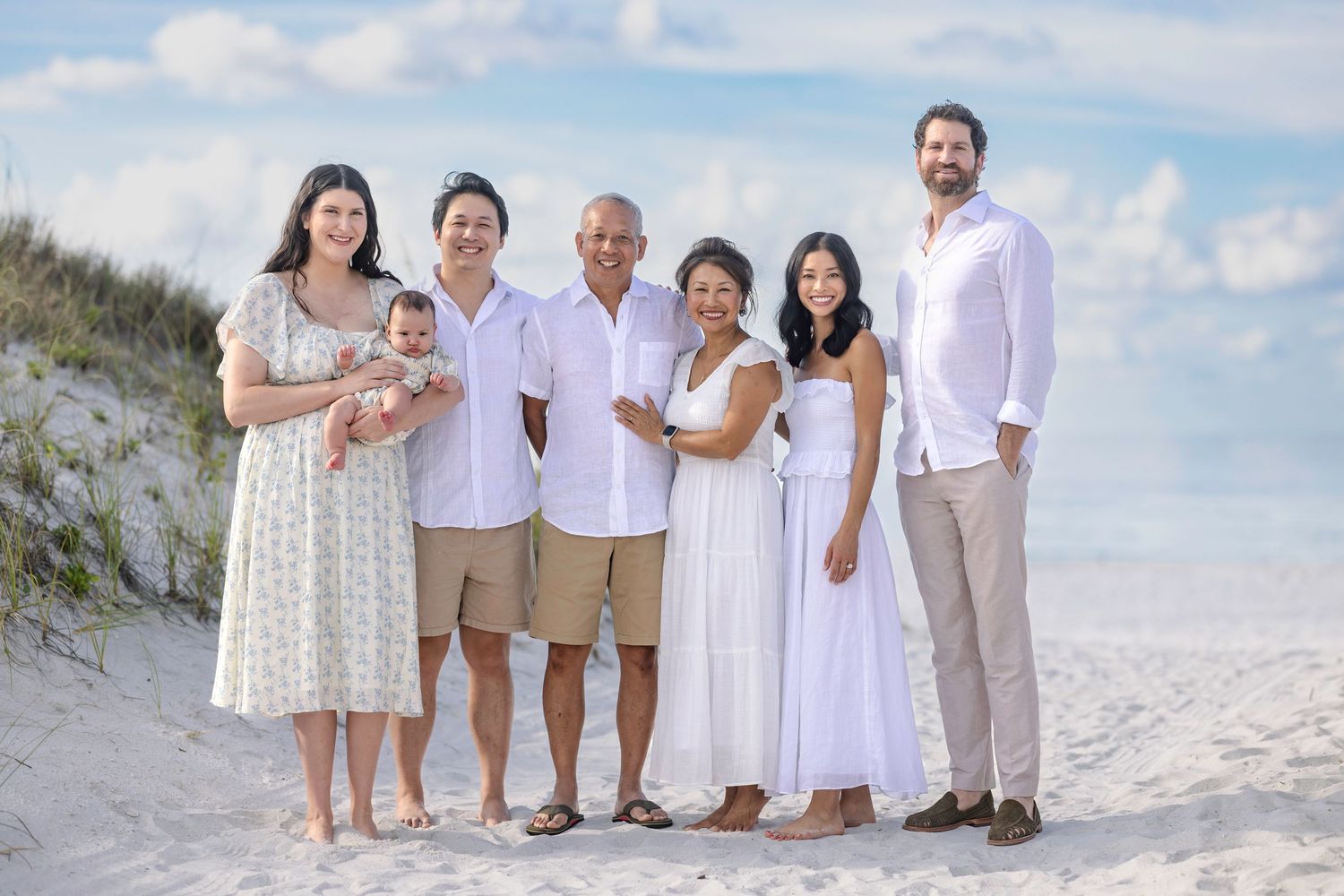 Multi-generational family portrait on sandy beach, seven members in white and neutral outfits, smiling outdoors.