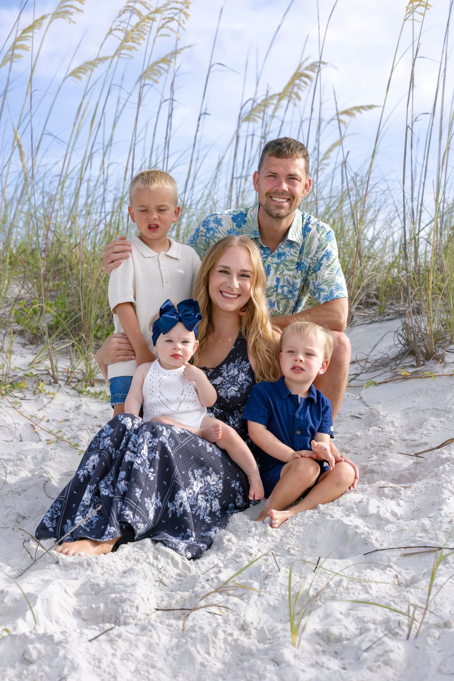Family of five posing on white sand beach with sea oats, mom in navy floral dress holding baby girl with blue bow.