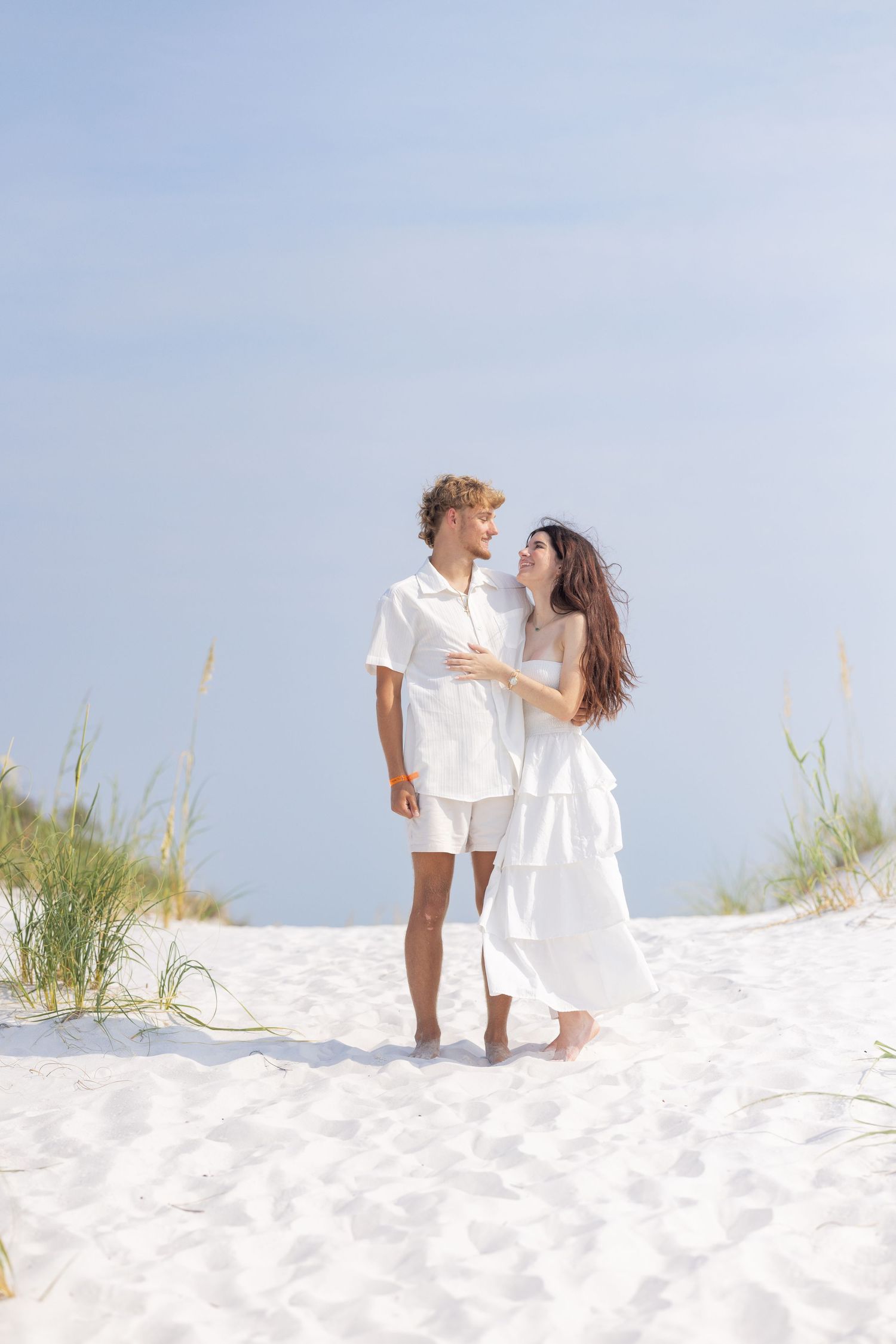 A couple in white outfits walks barefoot on white sand dunes, gazing at each other near sea oats under a blue sky.