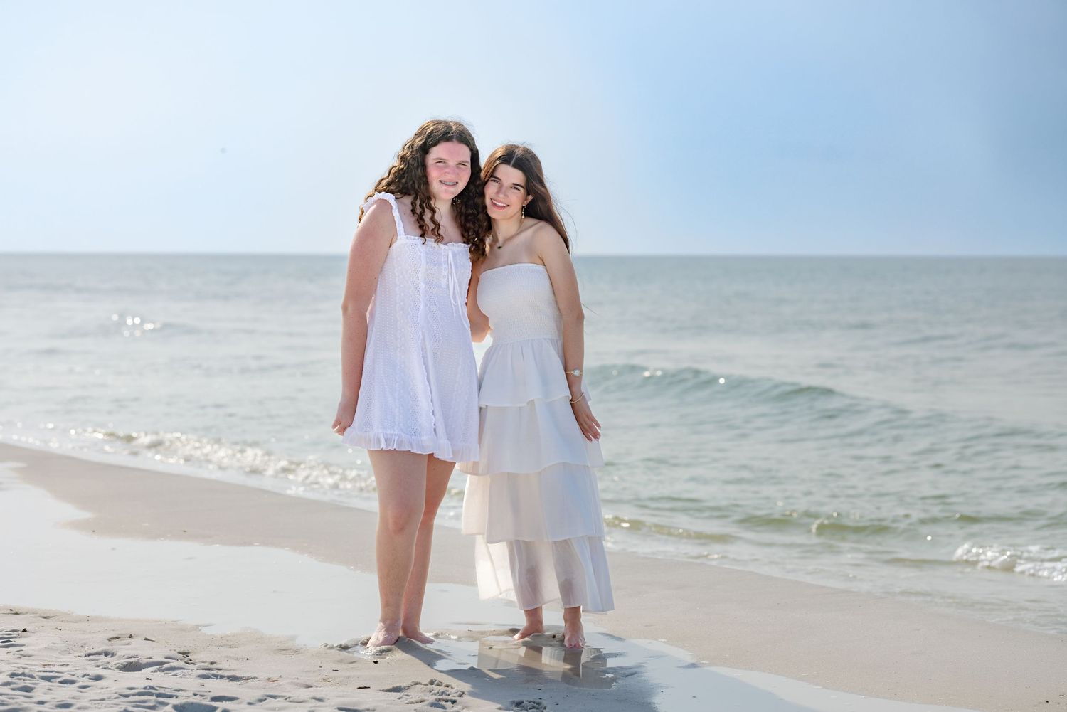 Two smiling women in white dresses posing together on a bright sandy beach by the ocean.