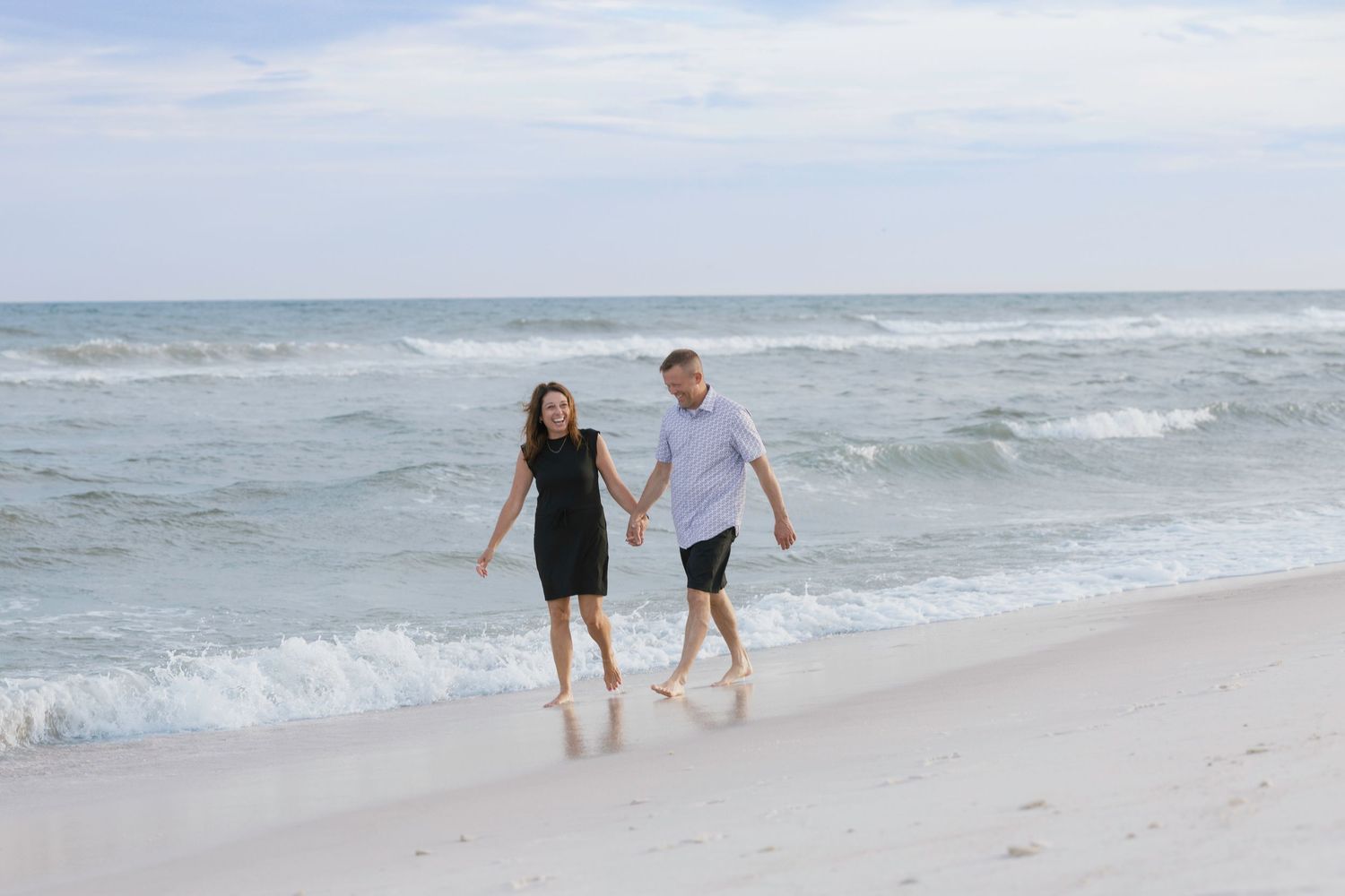 Couple holding hands walking barefoot on a sandy beach with ocean waves in the background.
