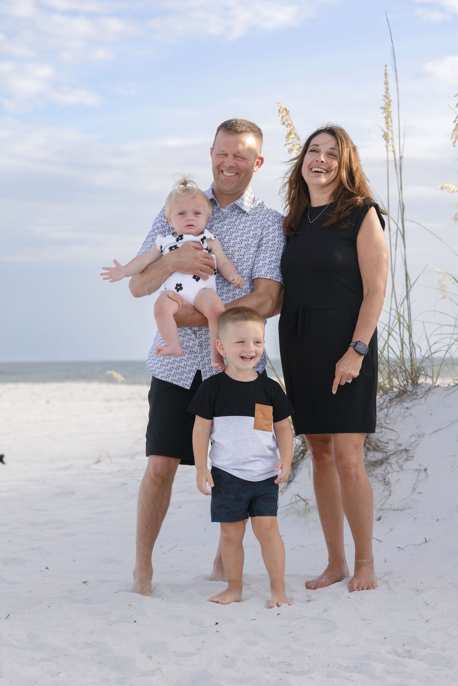 Happy family of four posing on white sand beach, parents holding baby girl with young boy standing in front.