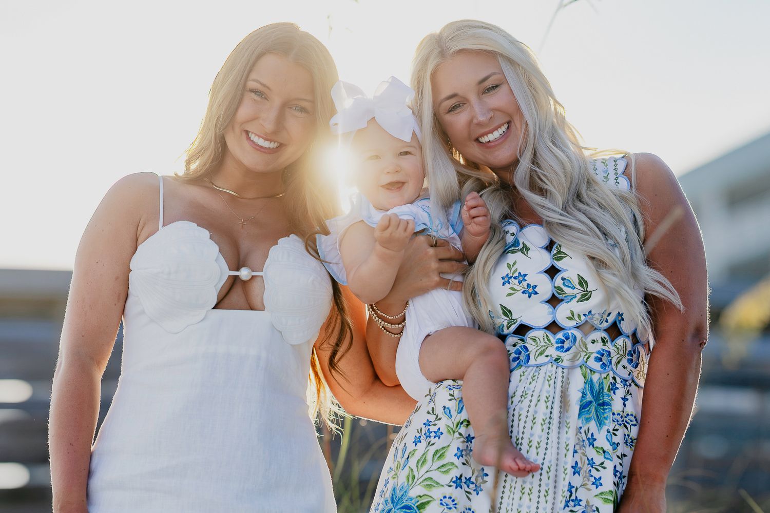 Two blonde women and a baby girl in a white bow pose outdoors in summer dresses with golden sunlight behind them.