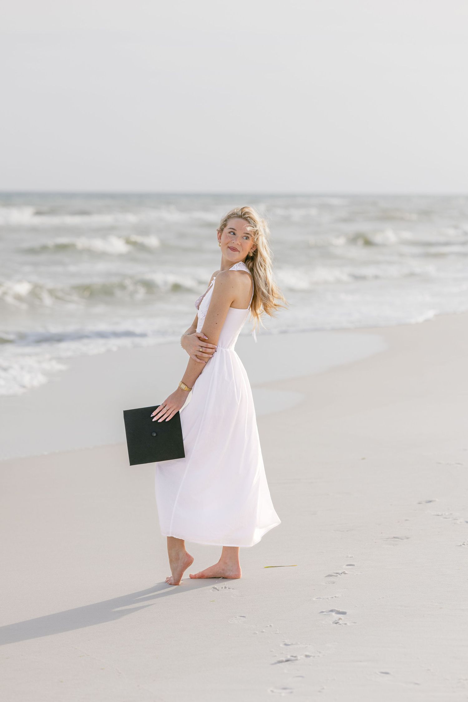 Young woman in white dress holding a book walks barefoot on a sandy beach with ocean waves behind her.