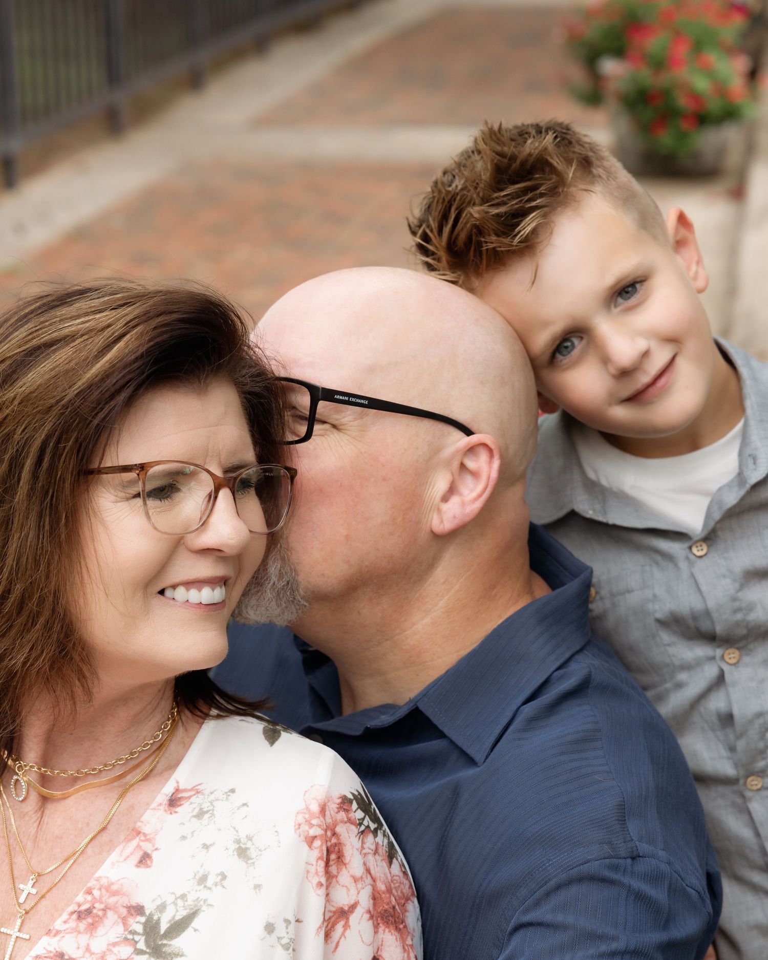 A bald man kisses a smiling woman while a young boy with blue eyes poses behind them outdoors.