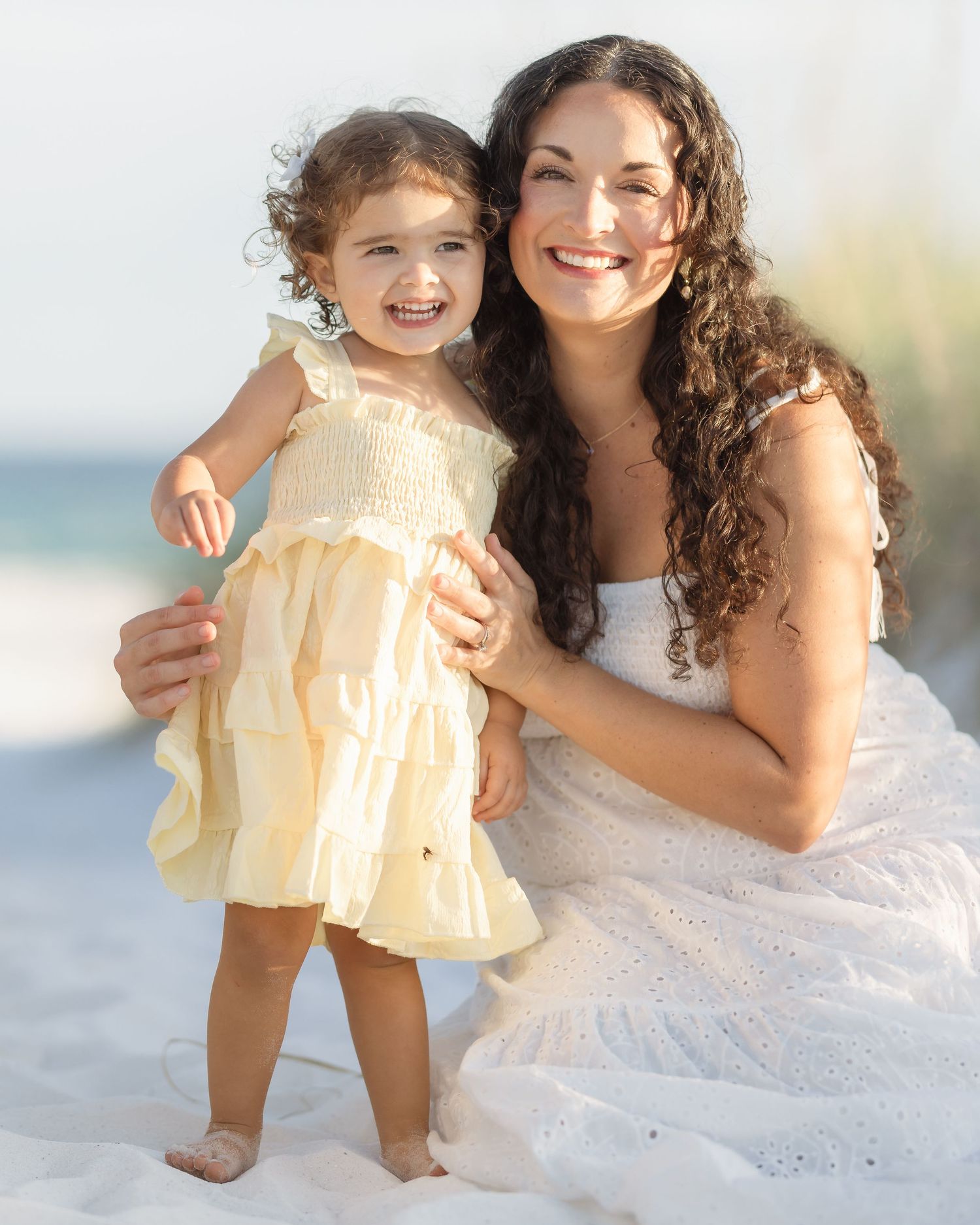 A smiling woman in white dress poses with a happy toddler girl in yellow dress on a sunny beach.