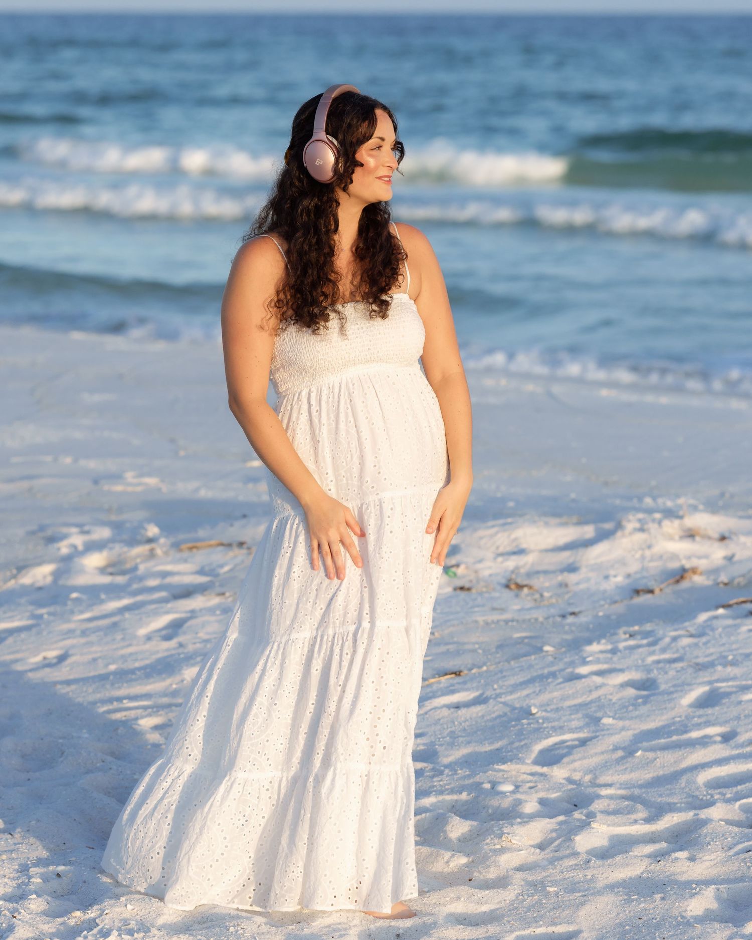 Woman in white maxi dress wearing pink headphones smiling on sunny ocean beach.