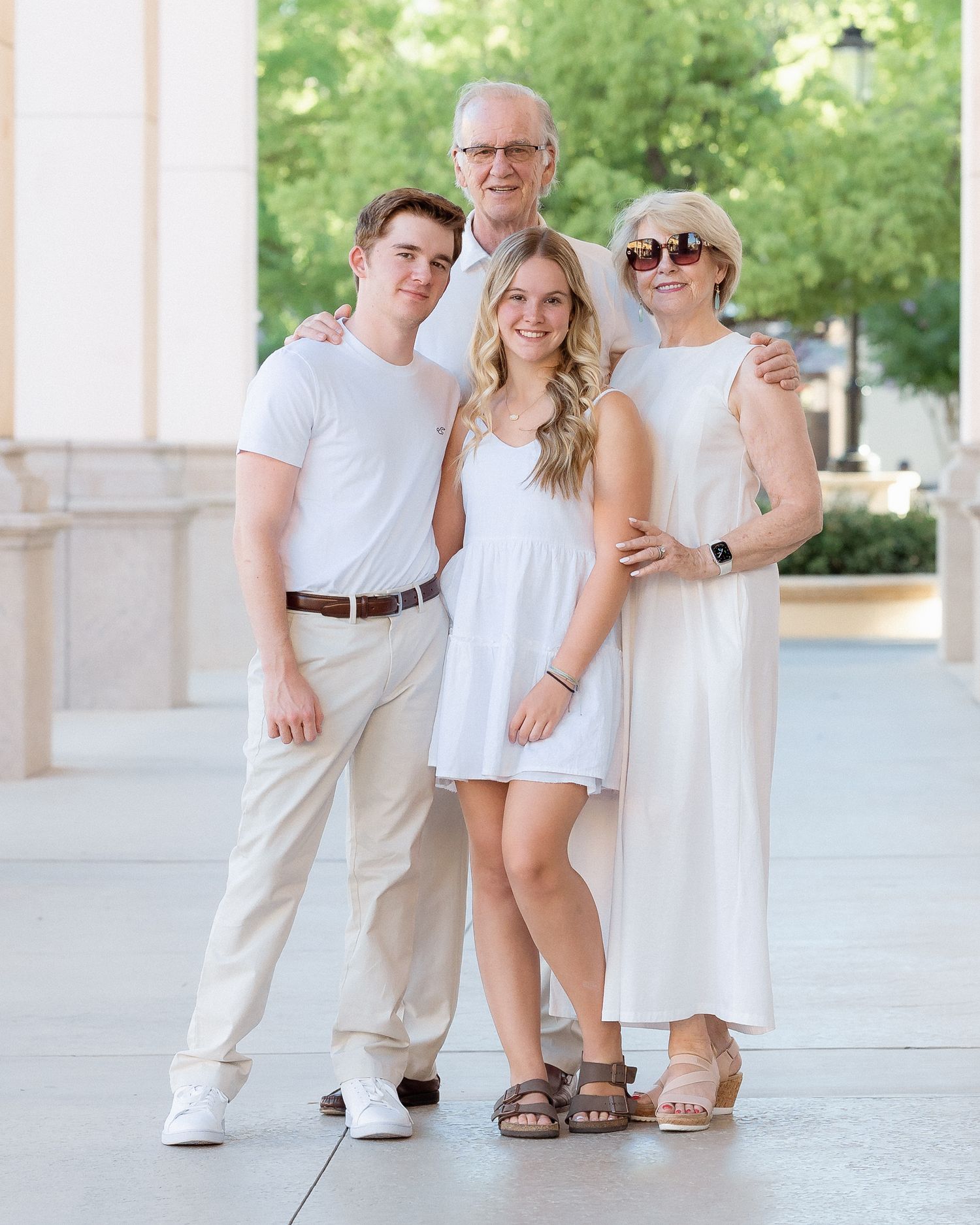 Family of four posing outdoors in coordinating white and beige outfits, smiling together in a sunny urban setting.