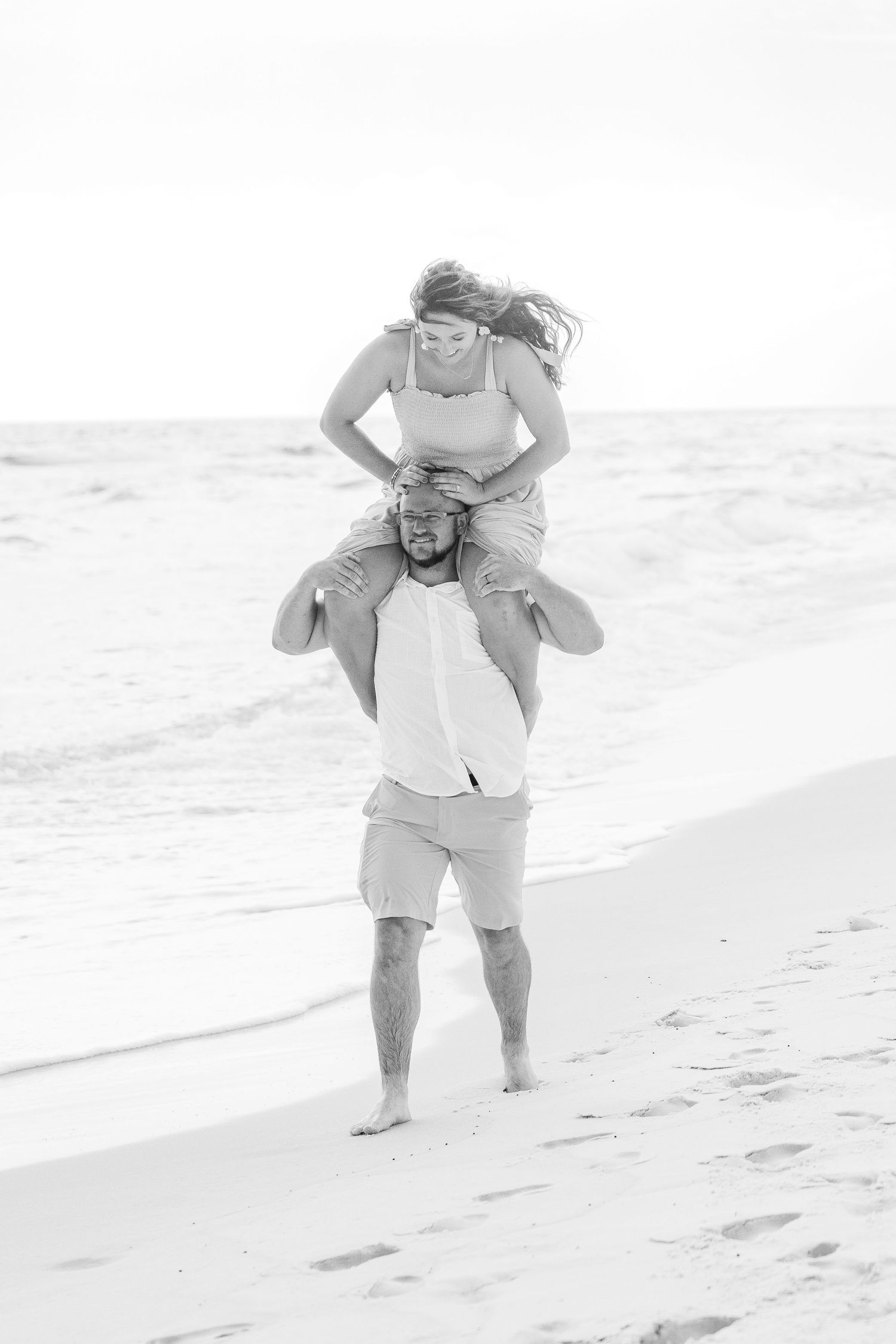 Black and white image of playful piggyback rides on empty beach shoreline.