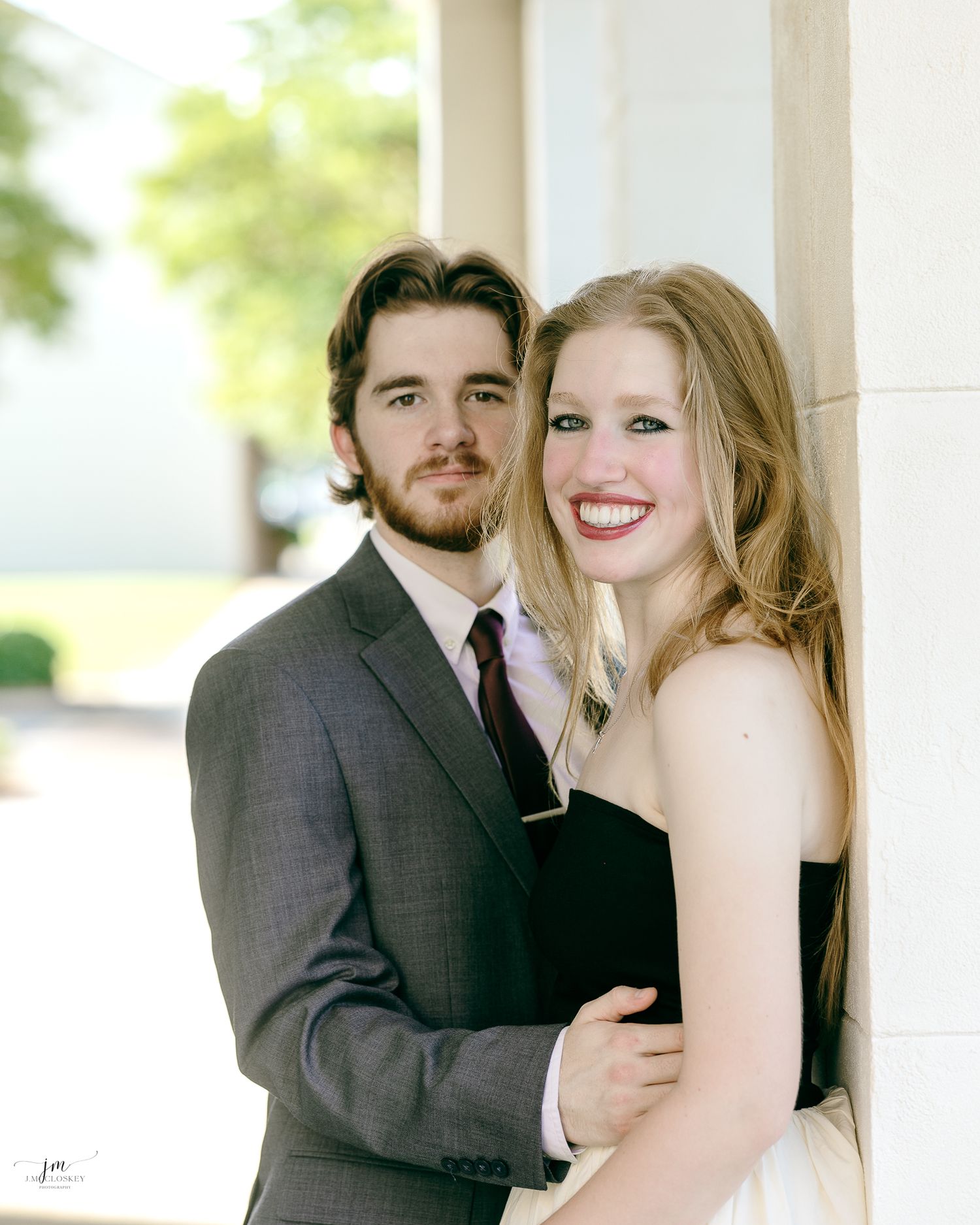 A couple embraces in formal wear against white columns during a sunny photoshoot.