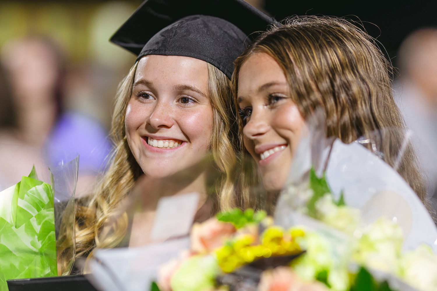 Two graduates smile while holding flower bouquets during graduation celebration.