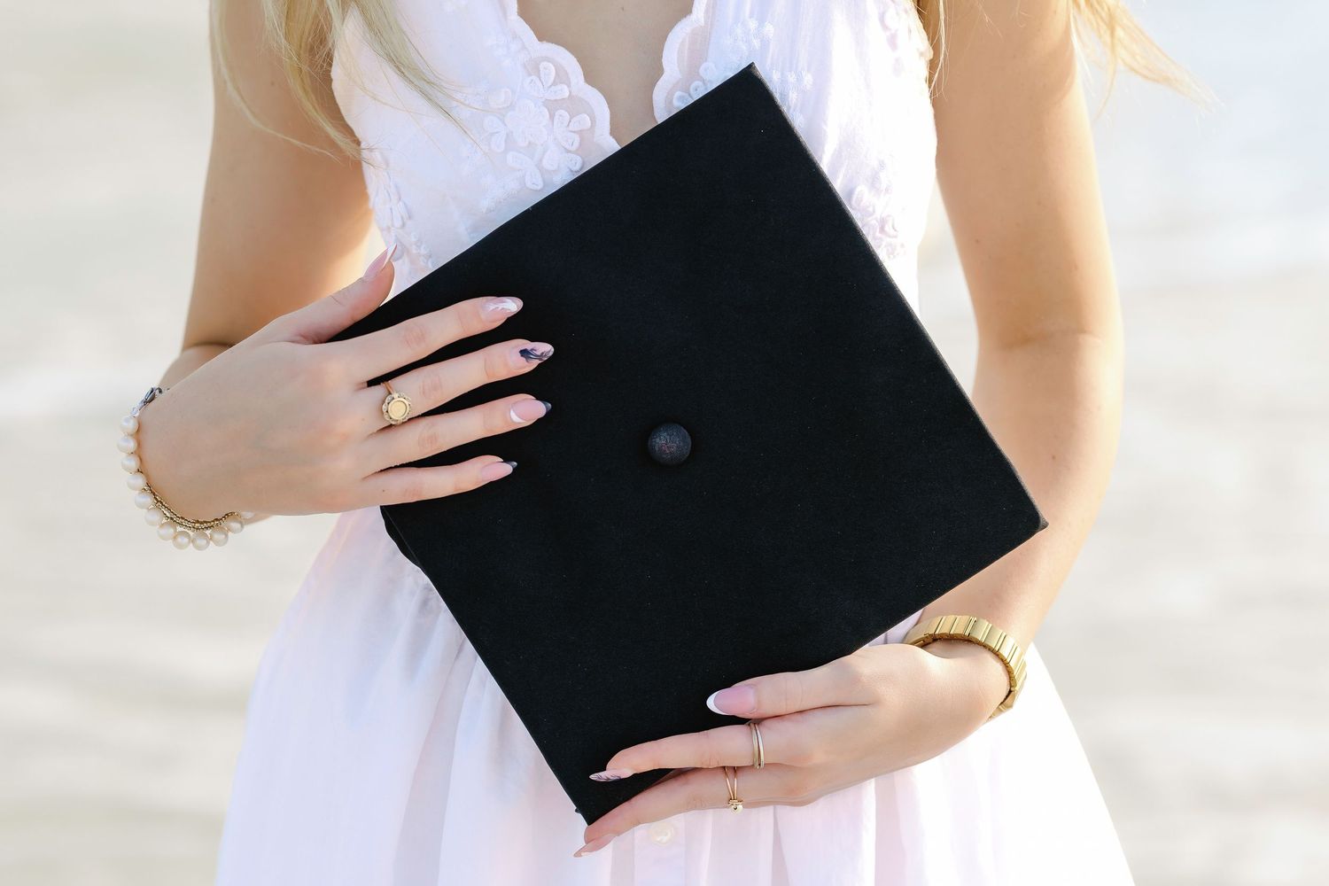 Graduate holding black diploma folder while wearing white dress against light background.
