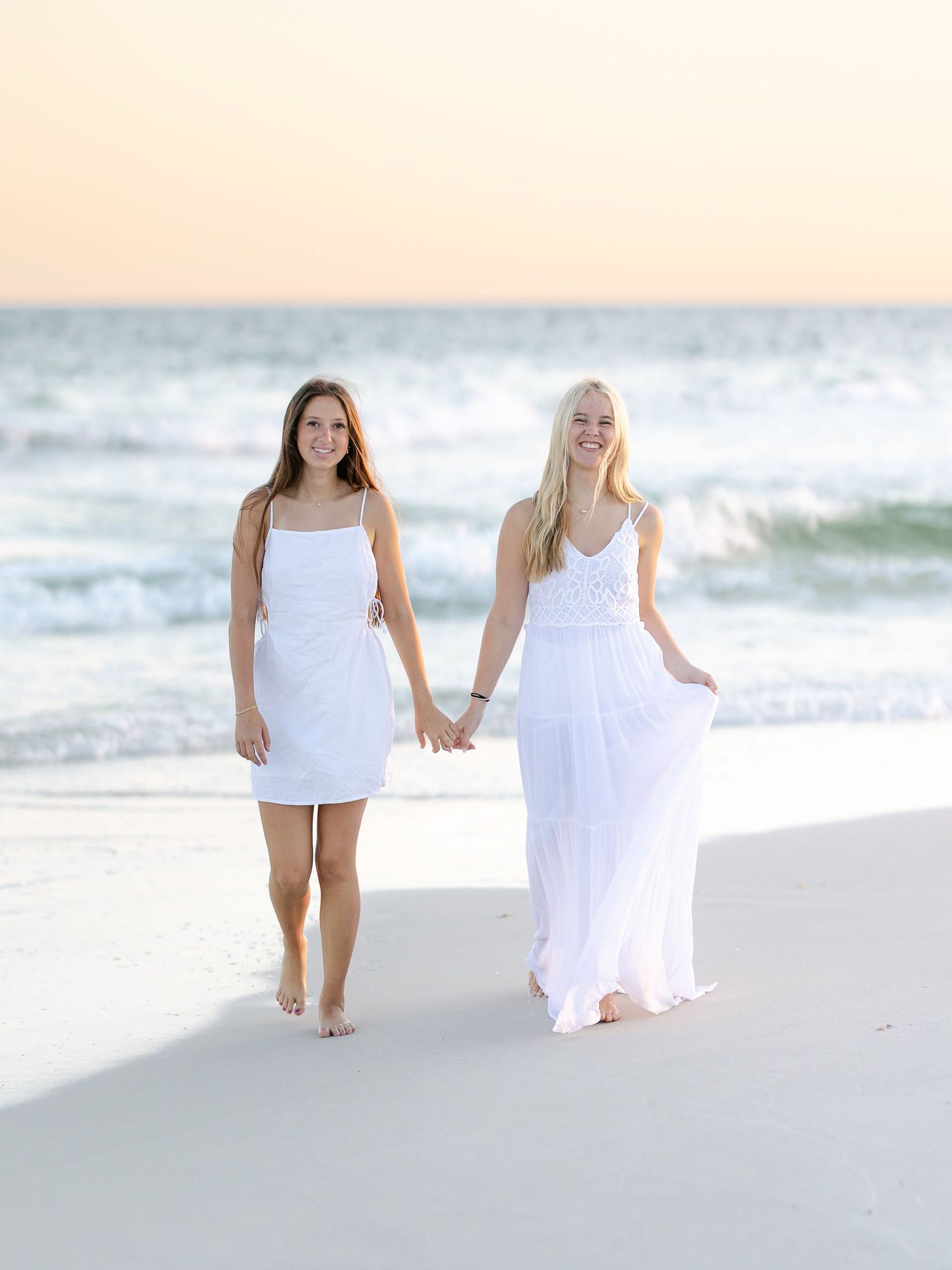 Two people walking hand in hand along beach shore in white dresses.
