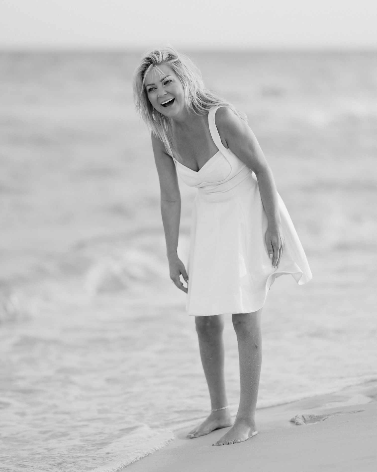 Black and white photo of laughing blonde woman in white dress on beach shoreline.