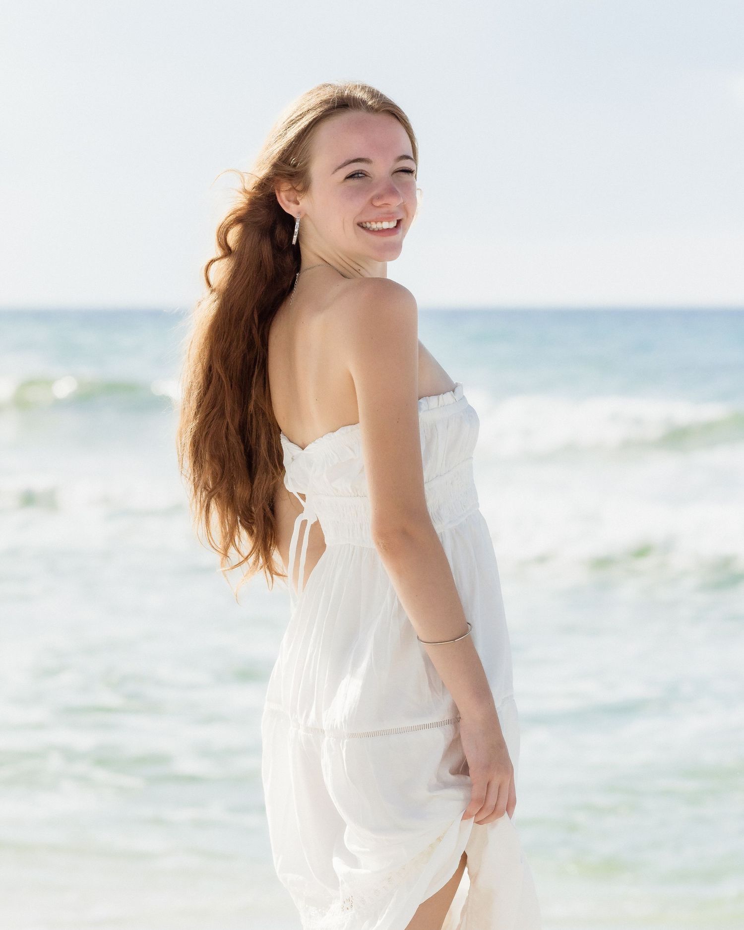 Young woman in white strapless dress smiling on a sunny beach with ocean waves in the background.