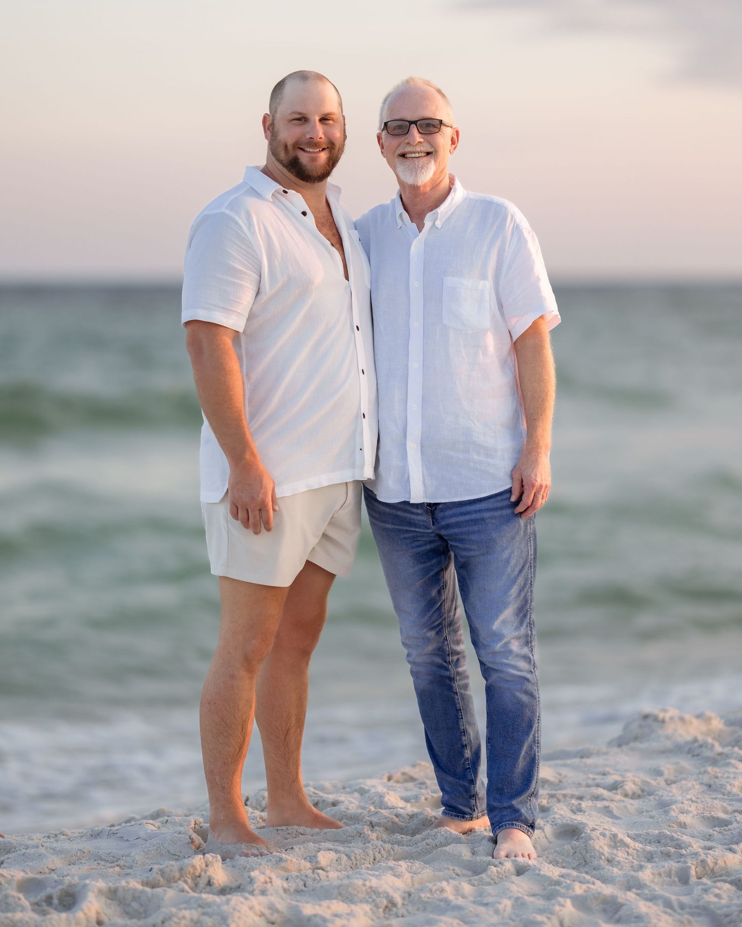 Two men in white shirts smile together on a sandy beach at sunset with calm ocean waves behind them.