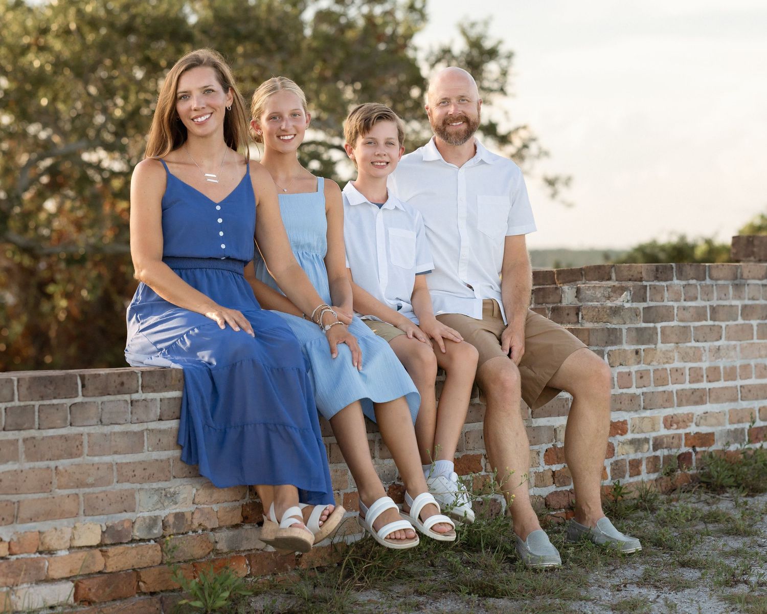 Family of four sitting on a brick wall outdoors, smiling and wearing coordinated blue and white summer attire.