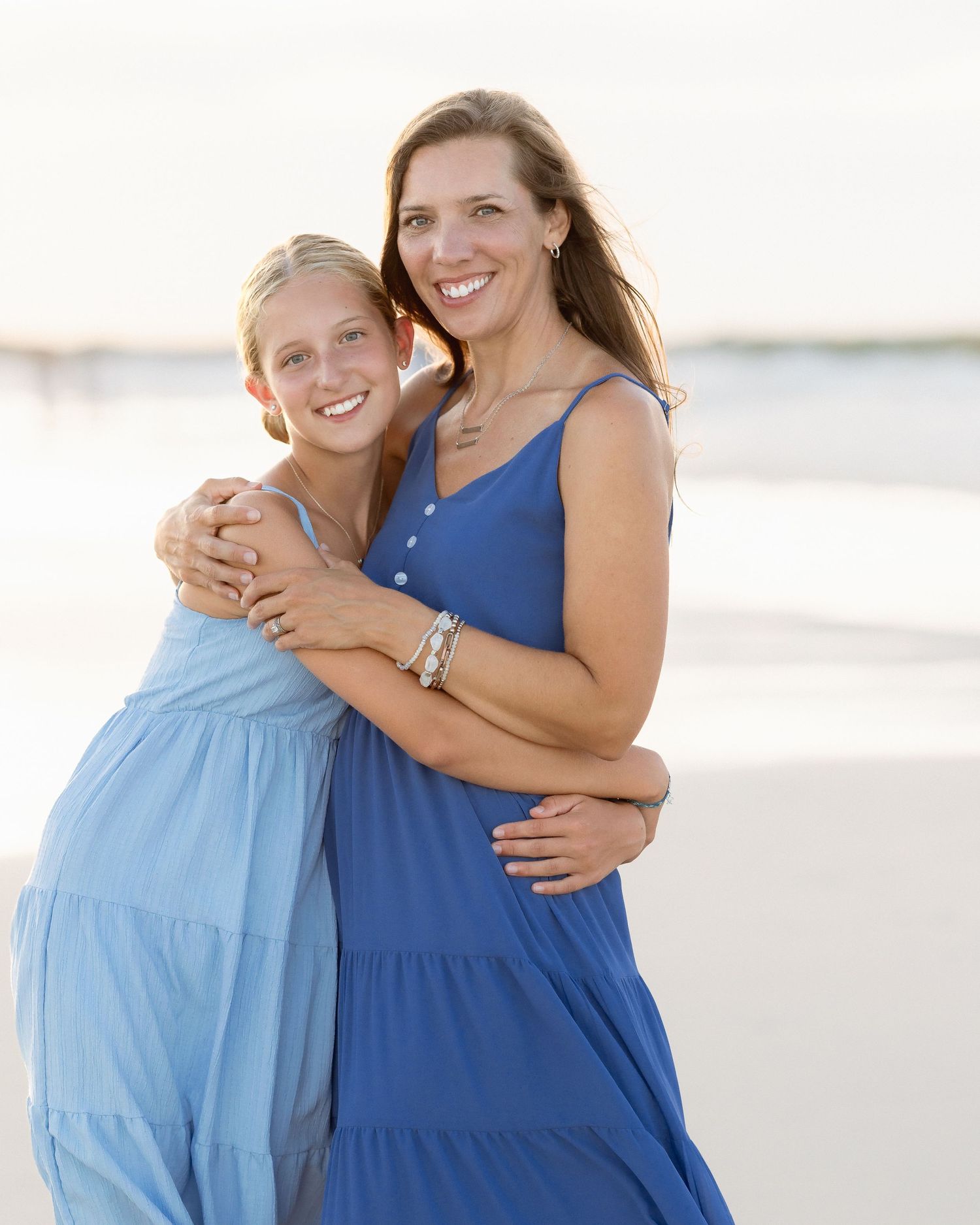 A mother and daughter in matching blue dresses smile and embrace on a sandy beach.