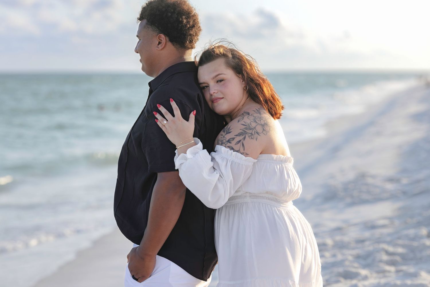 Couple embracing on a sandy beach at sunset, woman in white off-shoulder dress hugging man in black shirt.