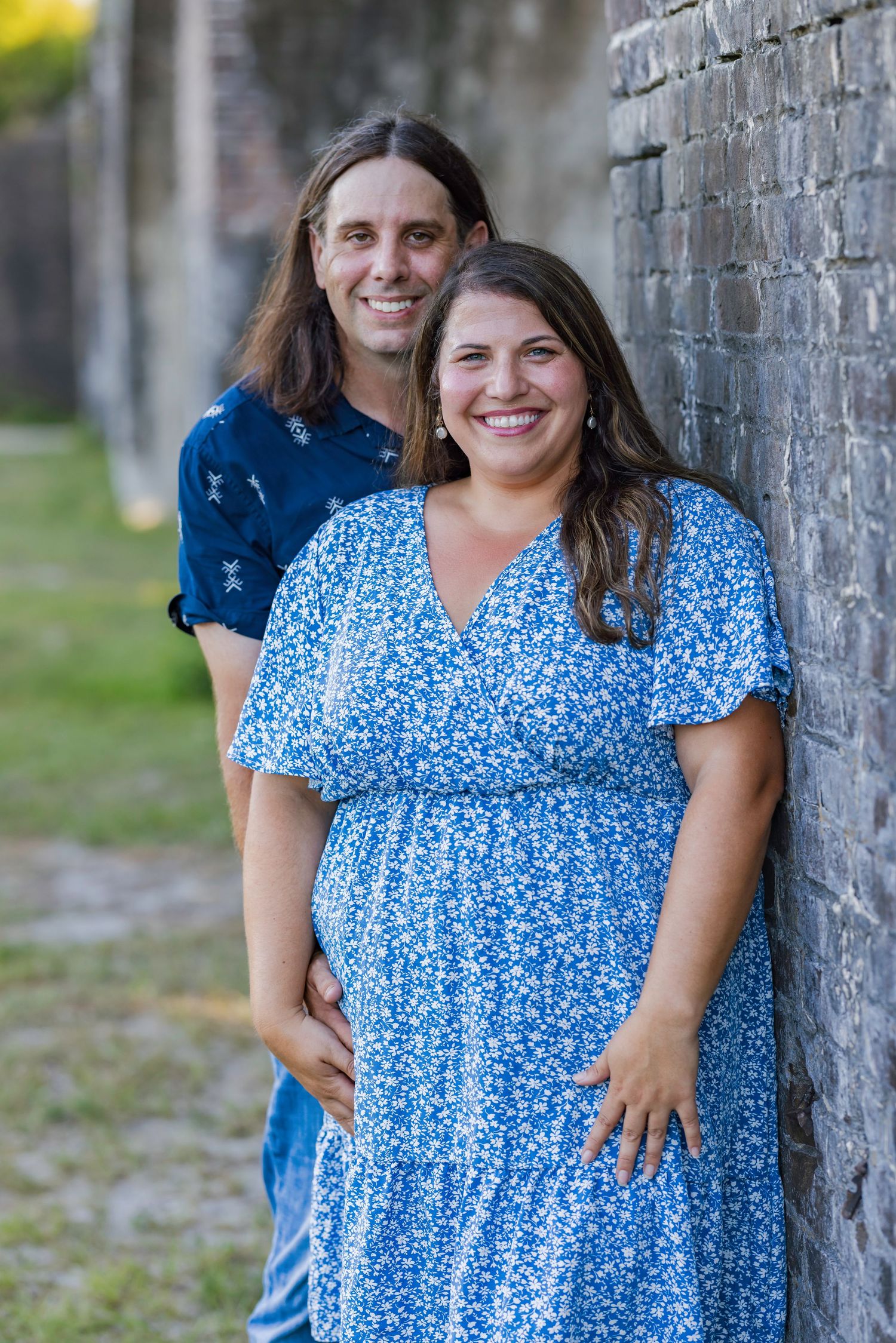 Smiling pregnant woman in blue floral dress poses with long-haired man against brick wall outdoors.