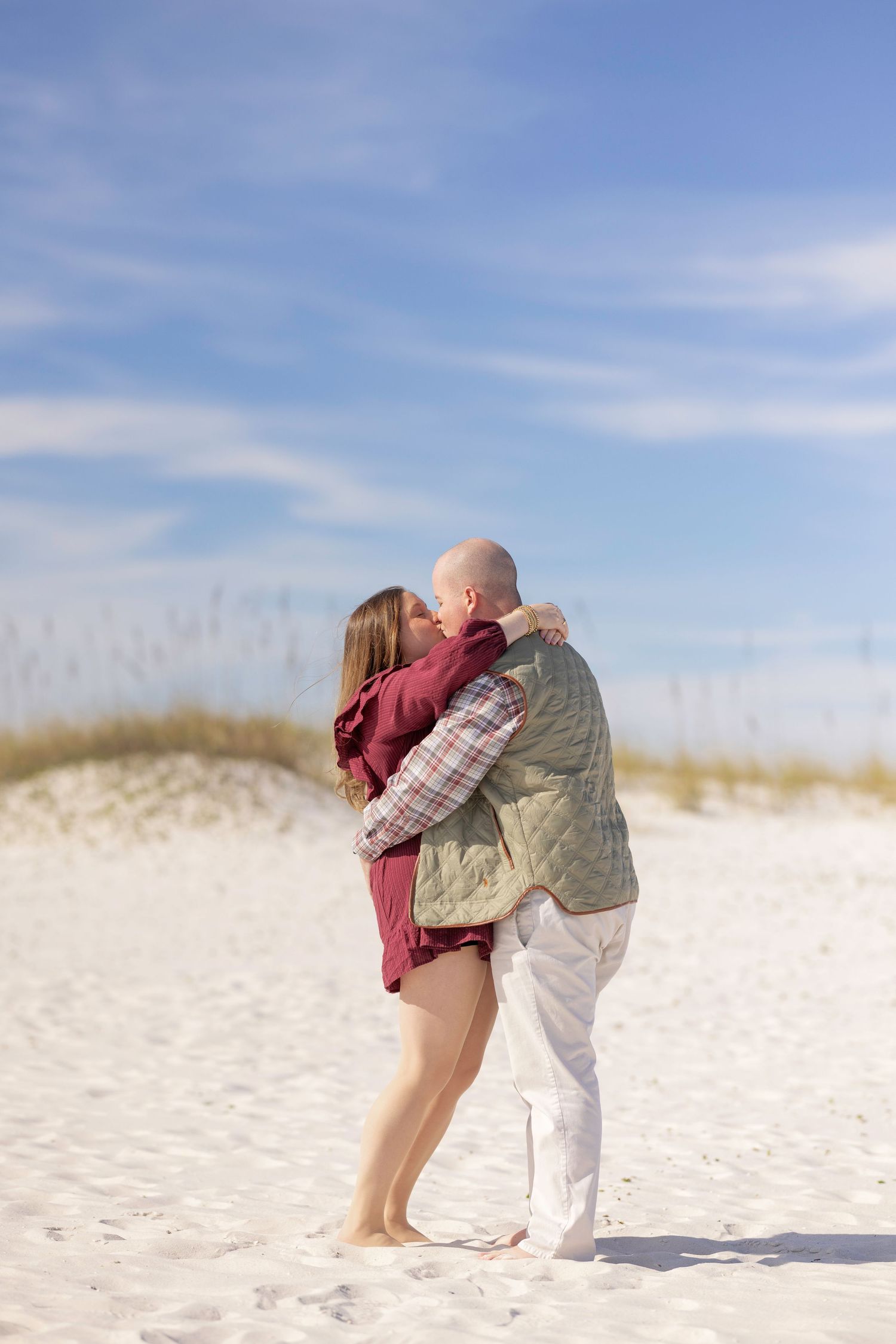 Couple embracing on white sand beach dunes under blue sky, woman in burgundy dress and man in quilted vest.