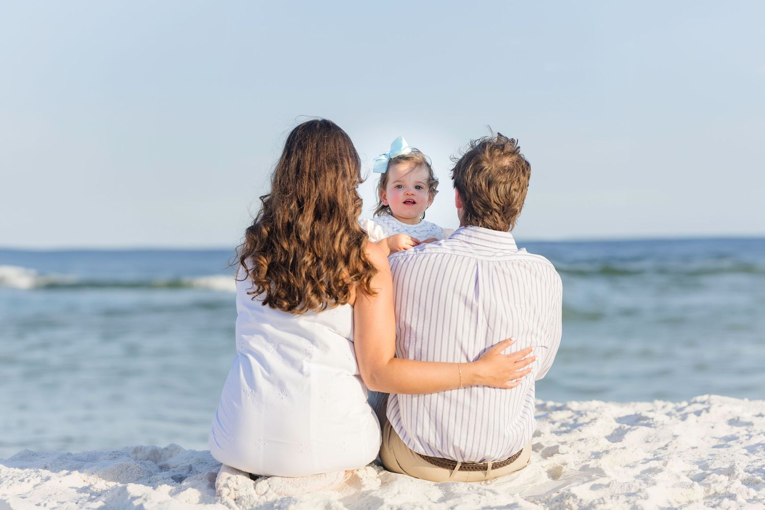 Family of three sitting on sandy beach facing the ocean with baby girl.