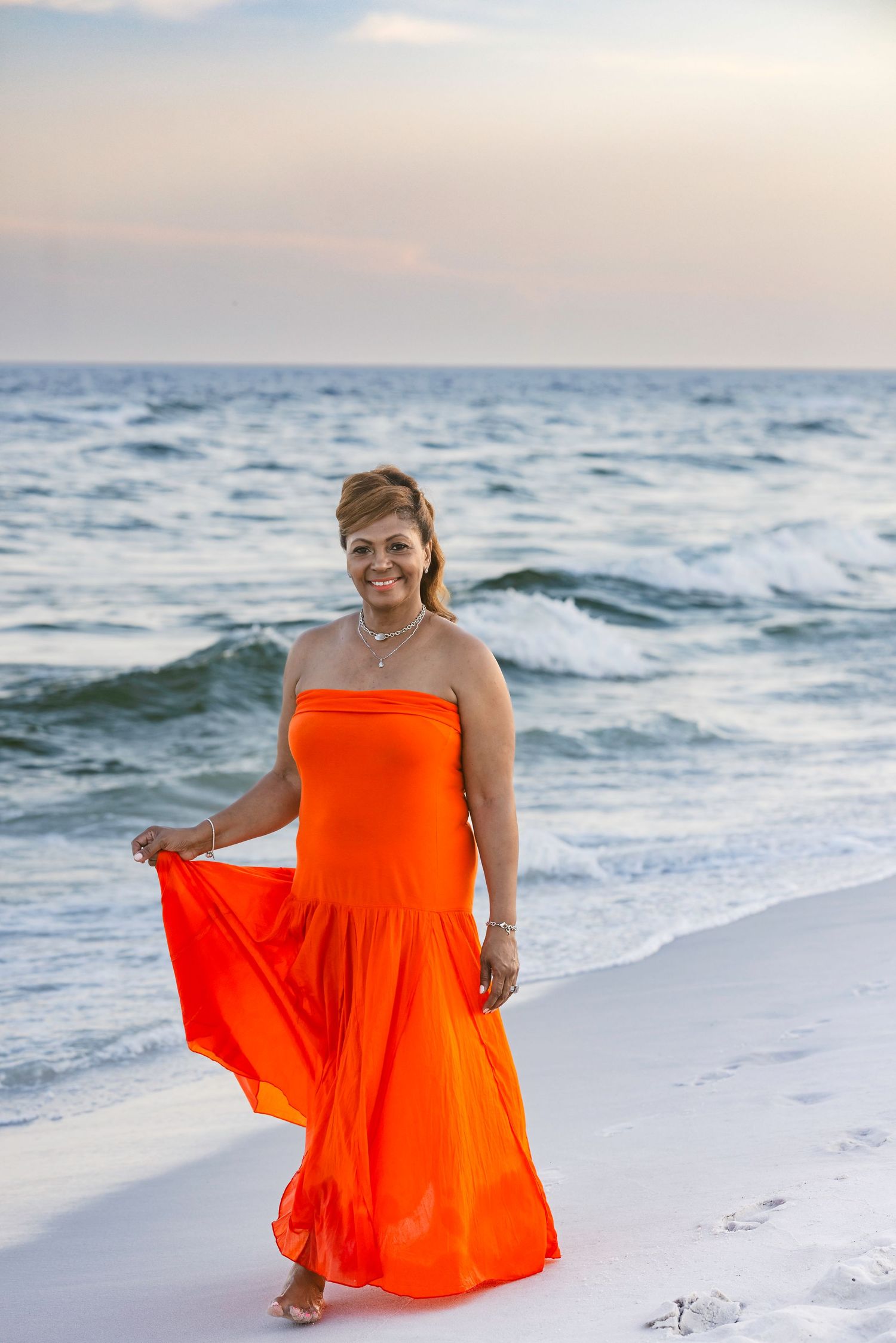 Smiling woman in flowing orange strapless maxi dress walking barefoot on white sandy beach at sunset.