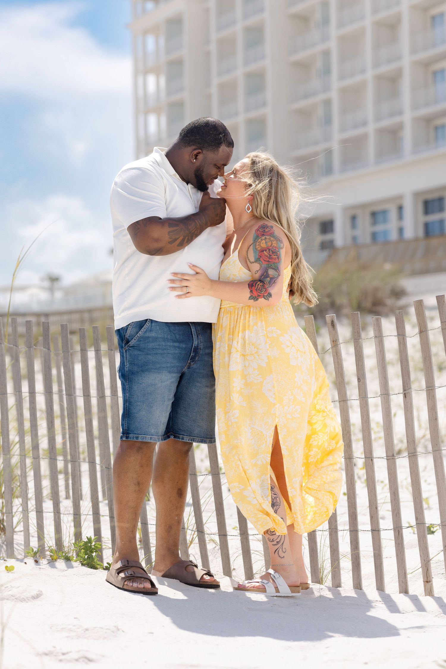 Interracial couple sharing a kiss on a sandy beach with a building in the background.