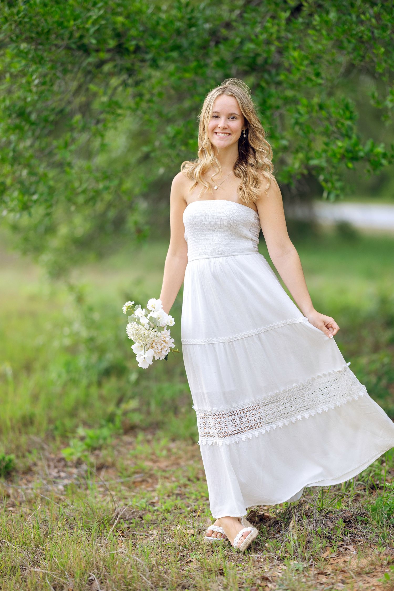 Young blonde woman in white strapless maxi dress holding white flowers, smiling outdoors in lush green setting.