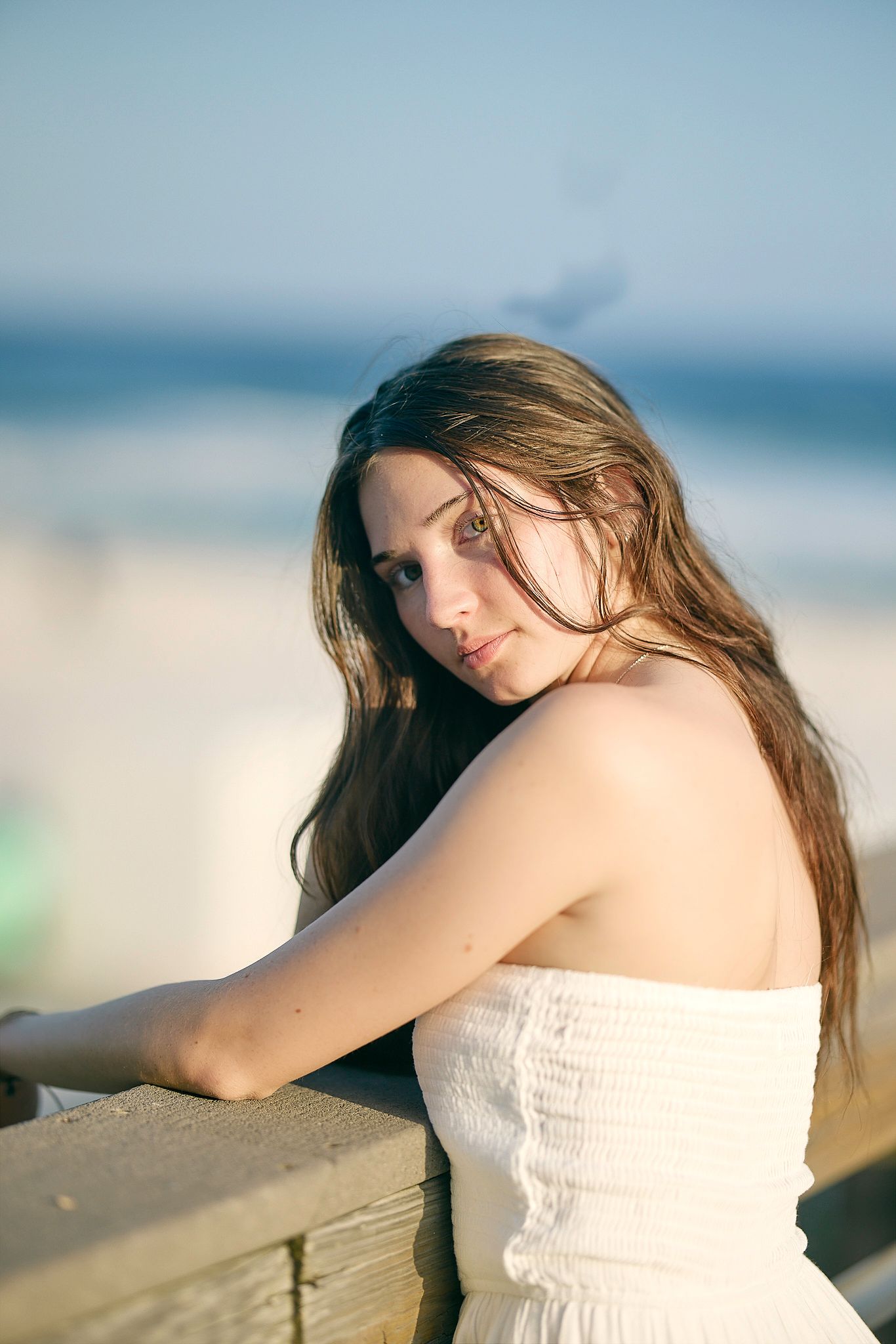 Young woman in white strapless dress leaning on wooden railing with ocean in the background.