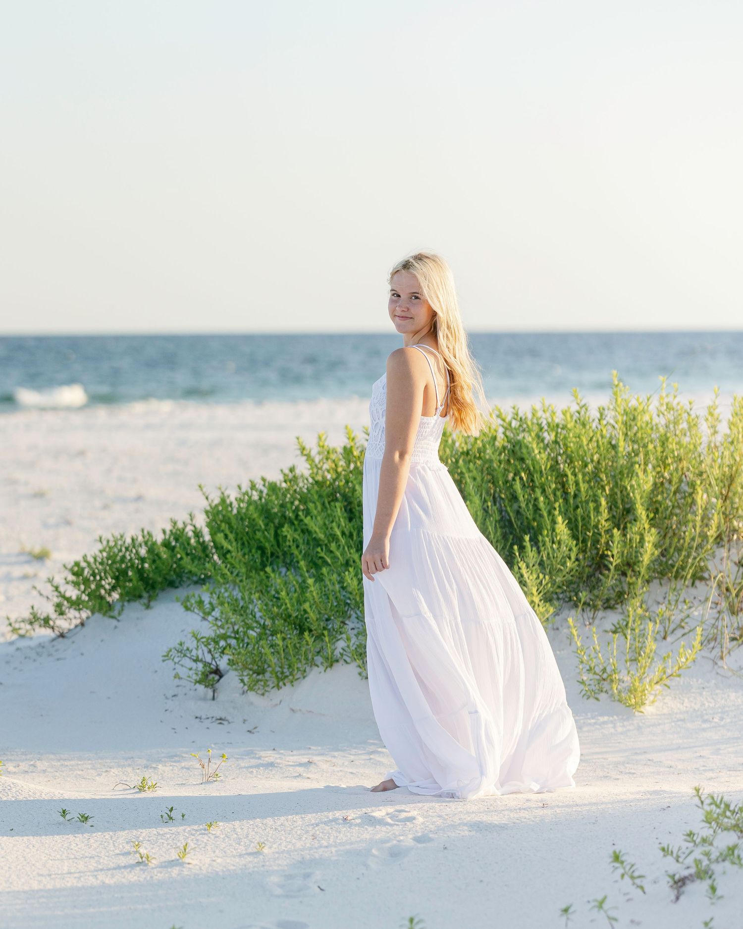 Young woman in flowing white dress standing on sandy beach with green dunes and ocean backdrop.
