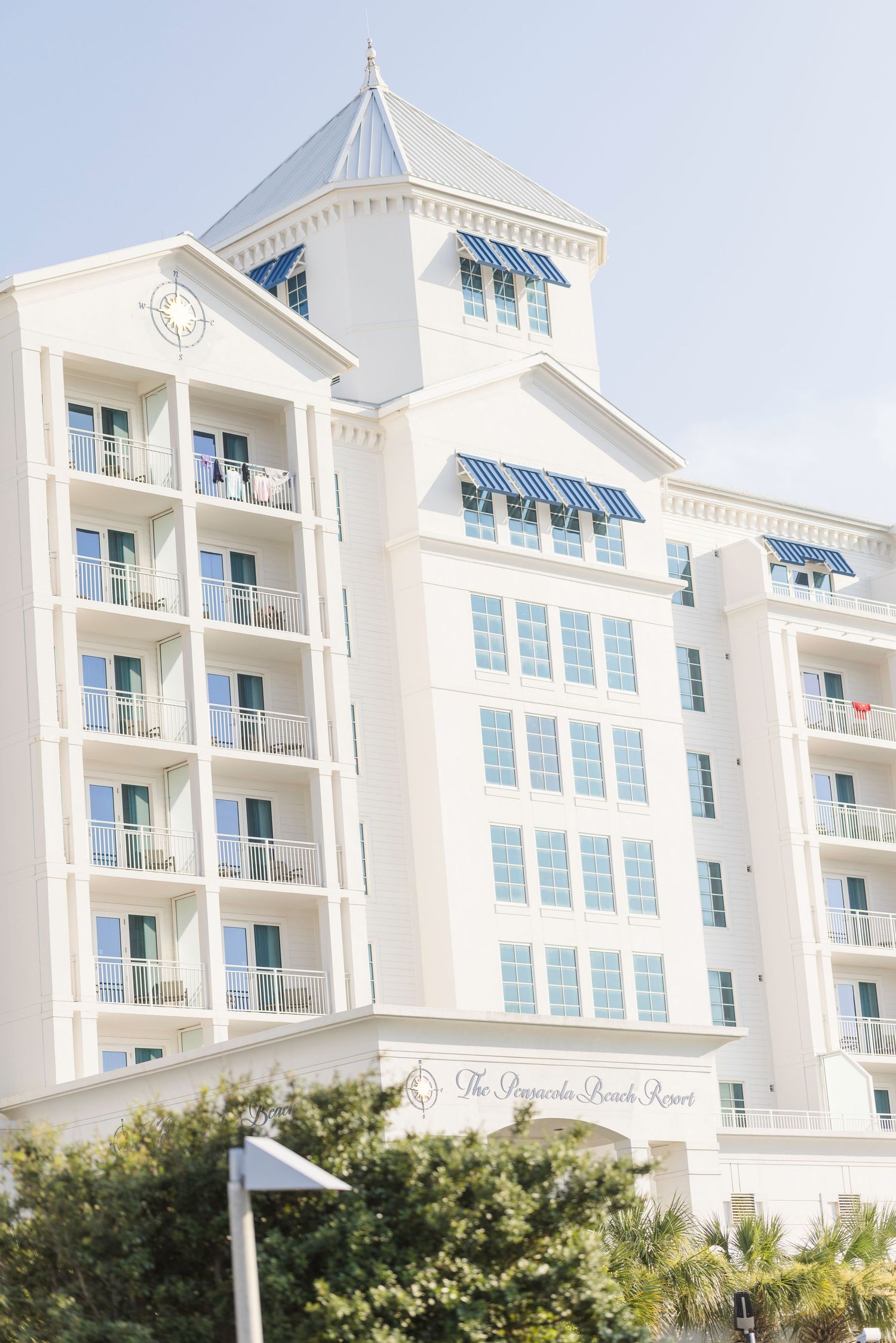 White multi-story Pensacola Beach Resort hotel with blue awnings, balconies, and a pointed tower against a clear sky.