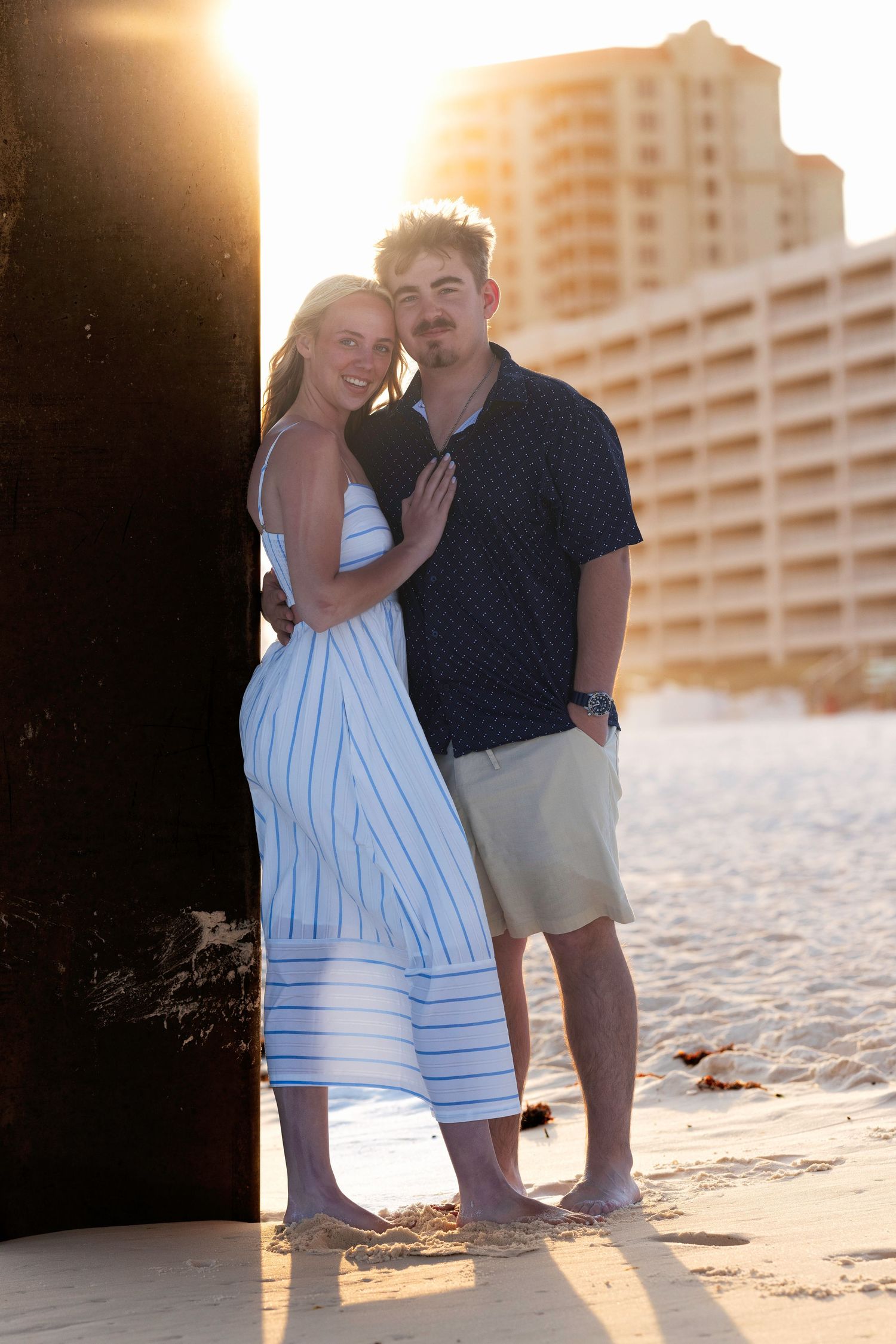 Couple embracing on sandy beach at golden sunset with coastal buildings in background.
