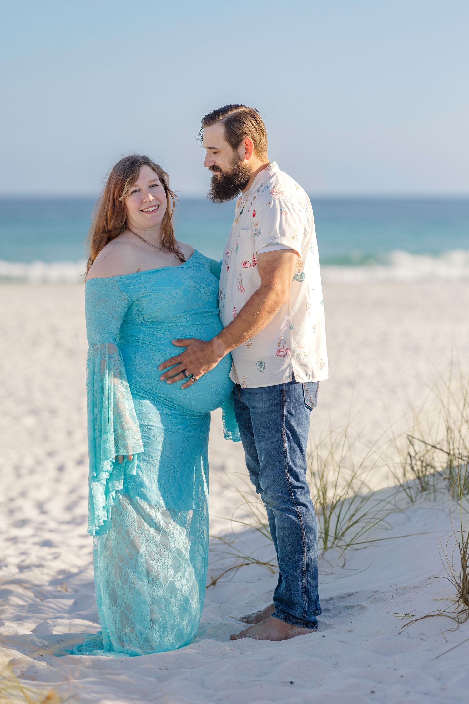 Couple sharing tender moment on beach, man holding pregnant woman's belly in teal gown.