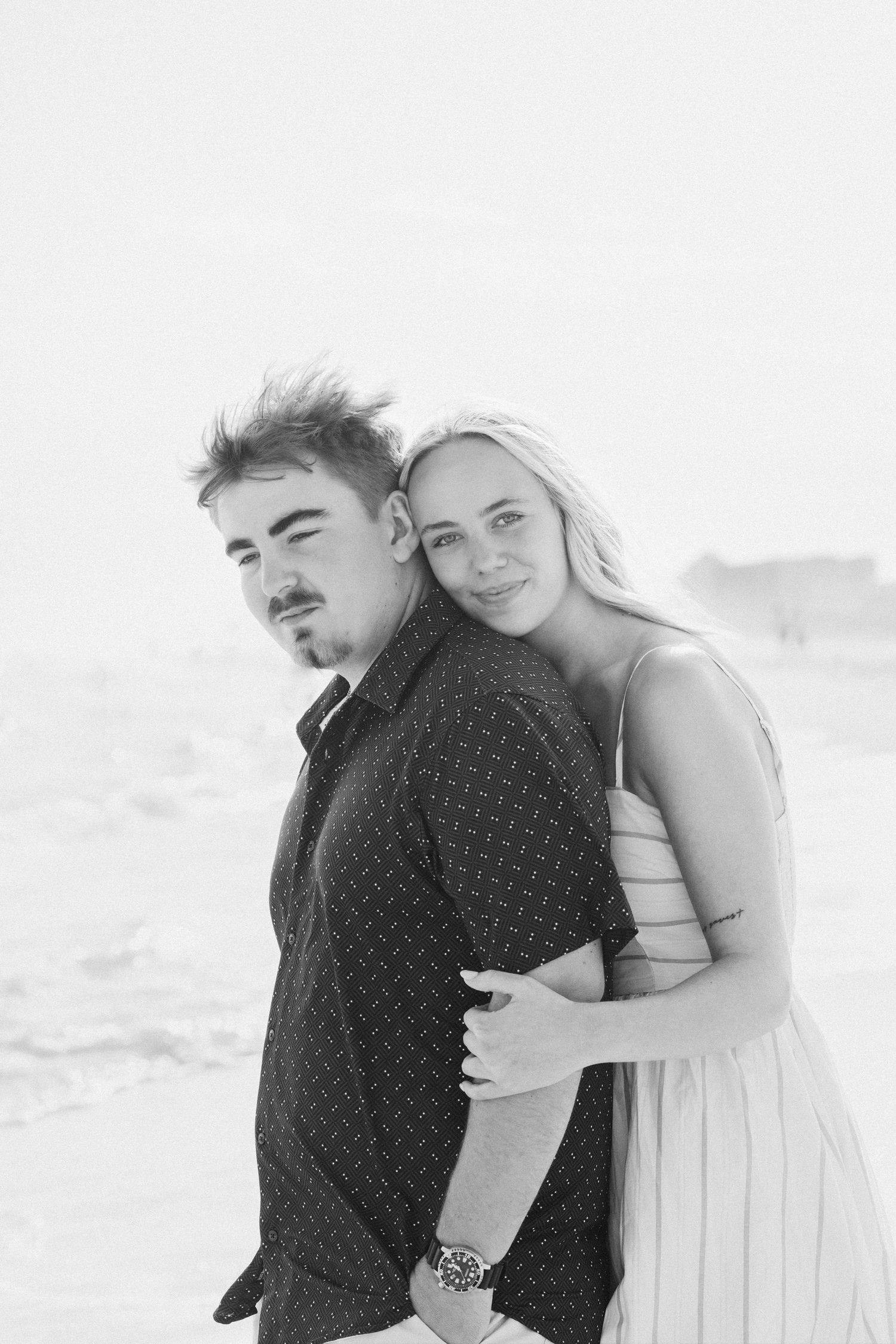 Black and white beach photo of a young couple embracing, woman in striped dress smiling behind man in dark shirt wearing a watch.