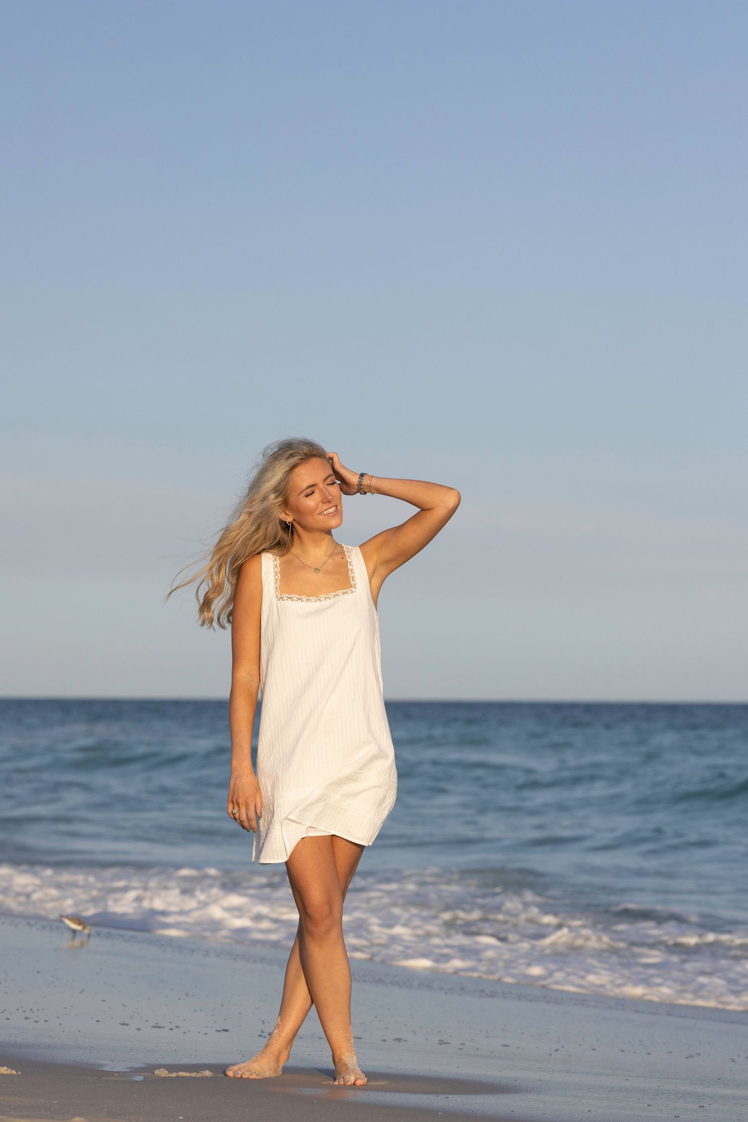 Smiling blonde woman in white dress walking barefoot on a sunny beach with ocean waves in the background.