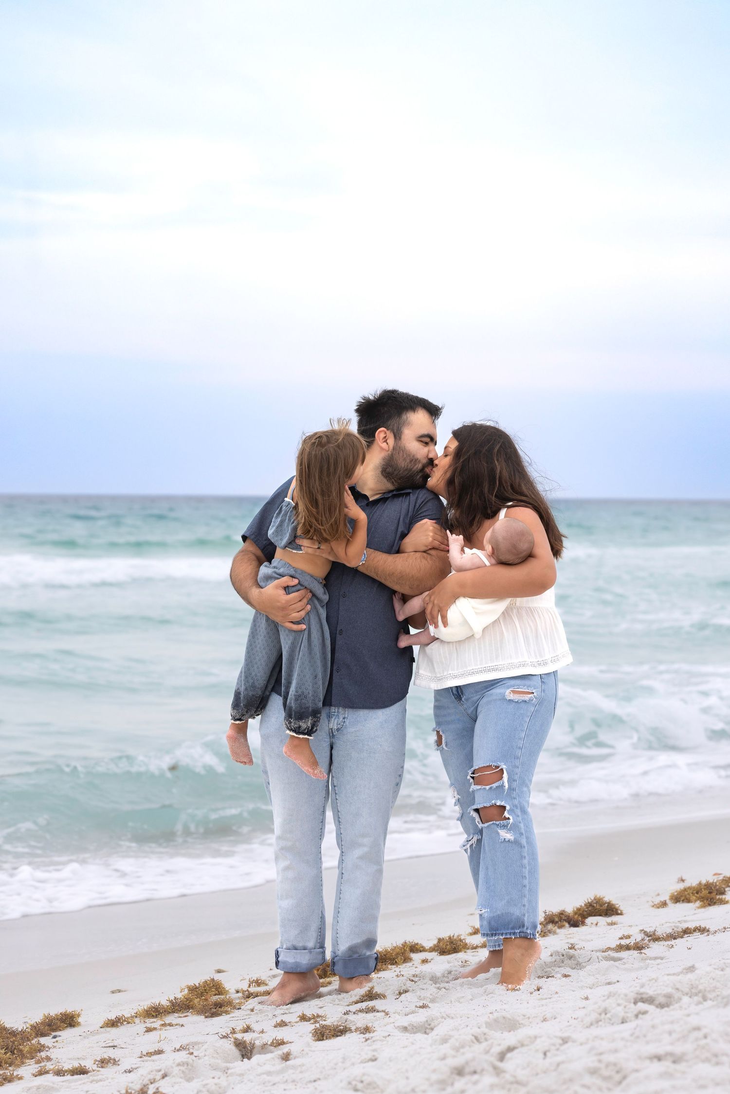 A family of four shares a tender kiss on a white sand beach, with parents holding two young children near ocean waves.