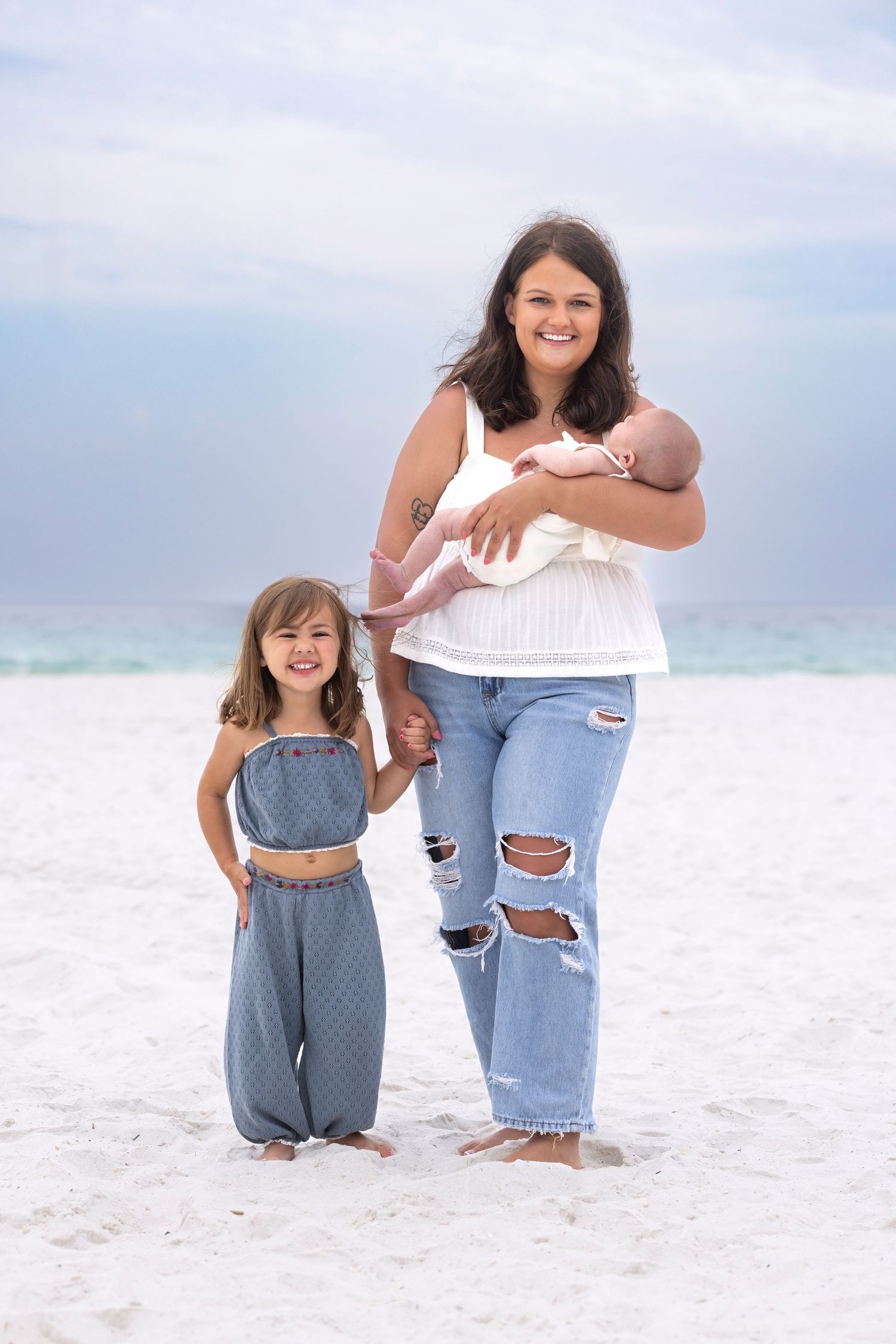A smiling mother holds a newborn while standing with her young daughter on a white sand beach near the ocean.