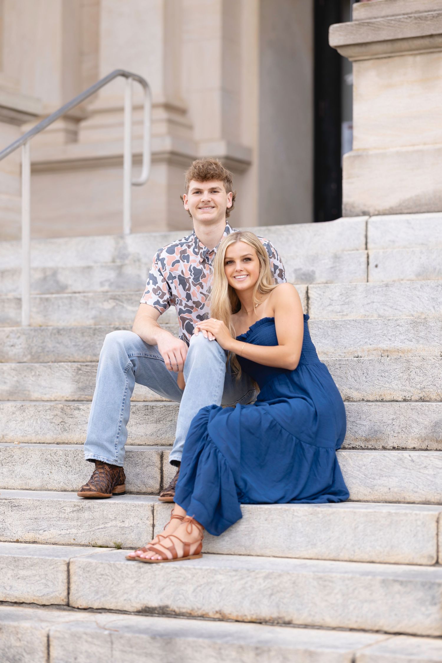 A couple in casual attire sits together on stone steps outside a building with white columns.