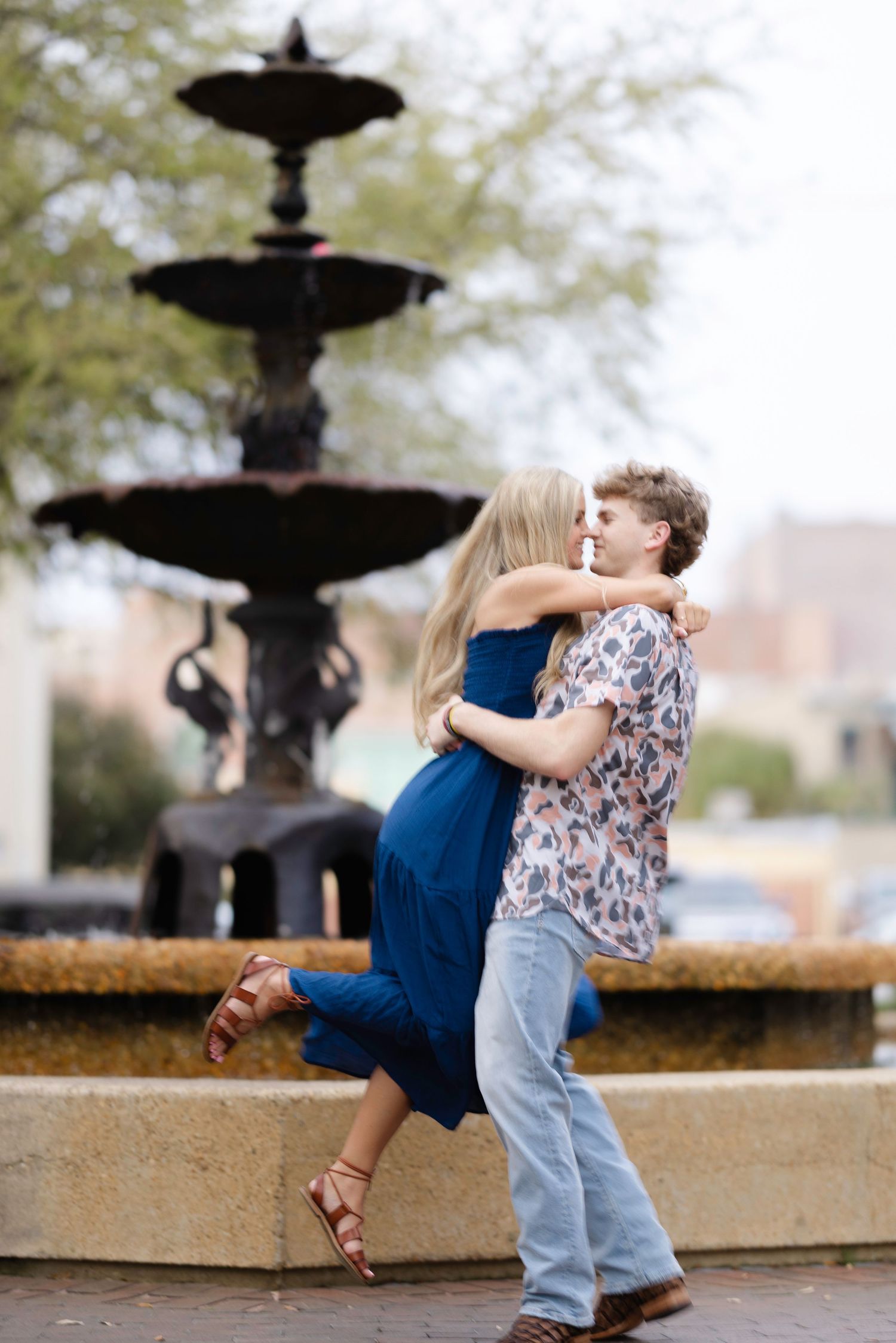 A couple embraces playfully by an ornate fountain in a romantic outdoor setting.