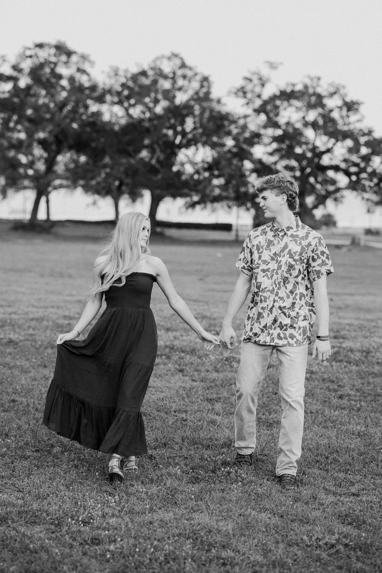 A romantic black and white photo of a couple holding hands while walking on grass with trees in the background.