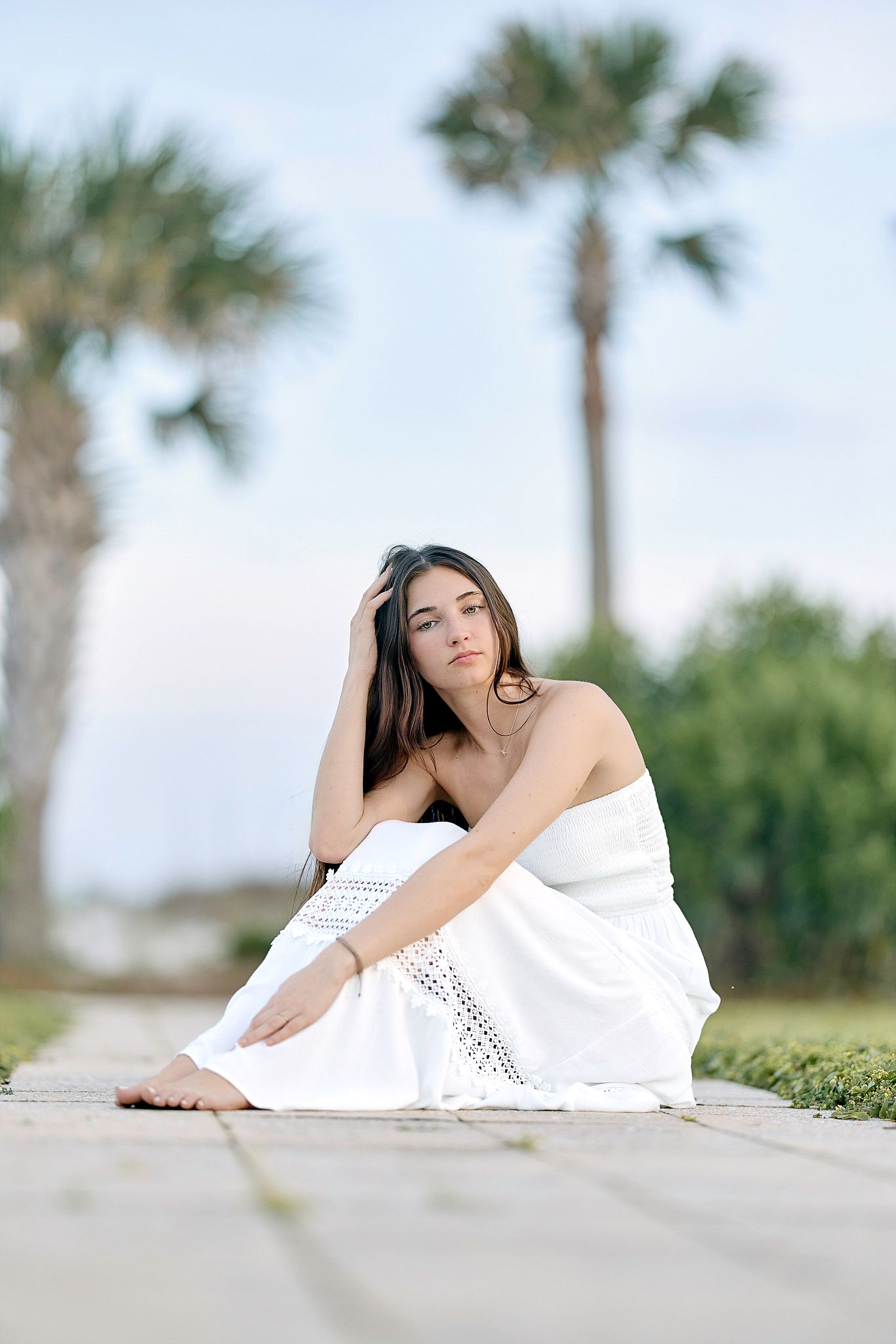 Person in white dress sitting on concrete path with palm trees in background on sunny day.