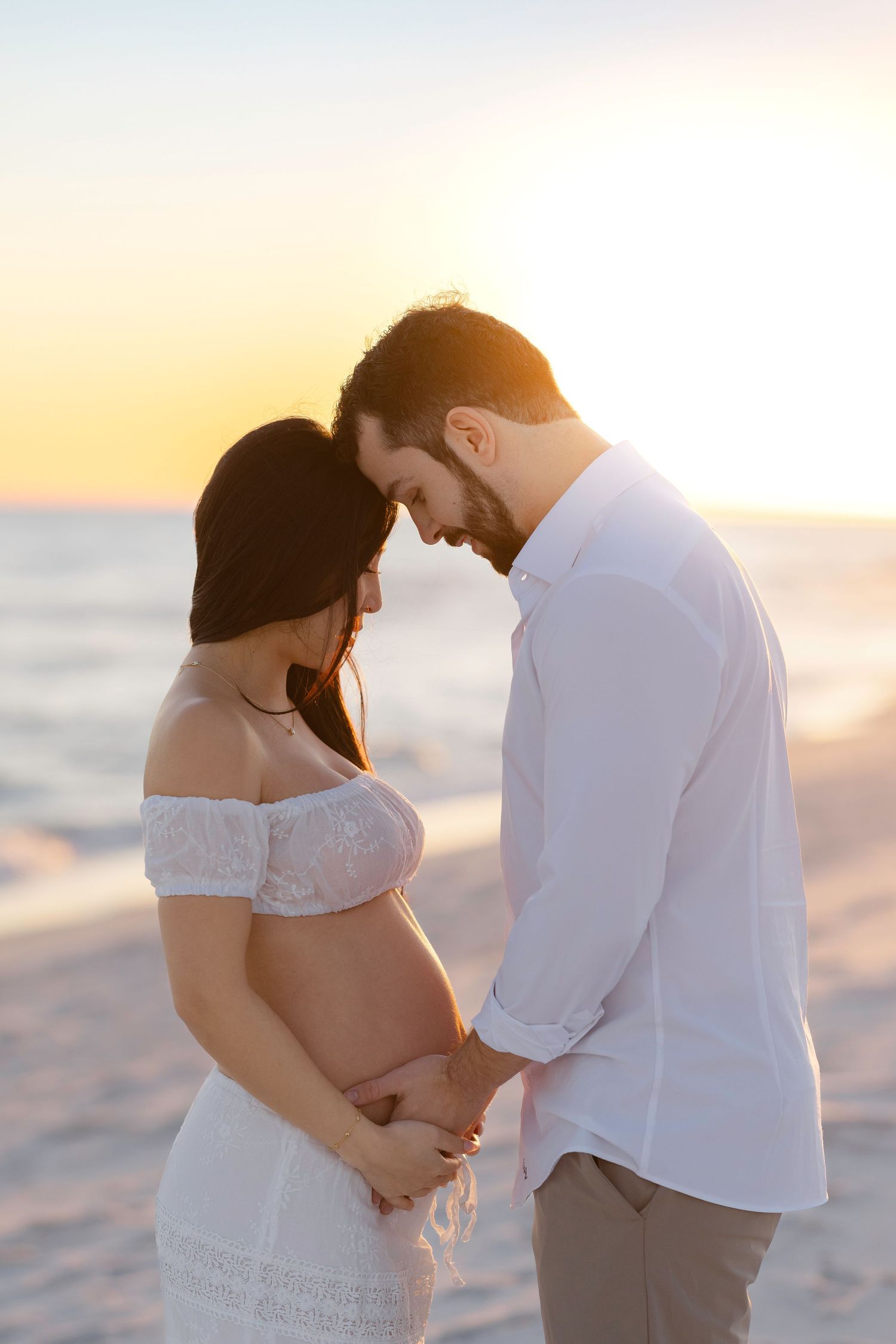 A romantic beach maternity photo session showing a couple embracing during sunset by the ocean.
