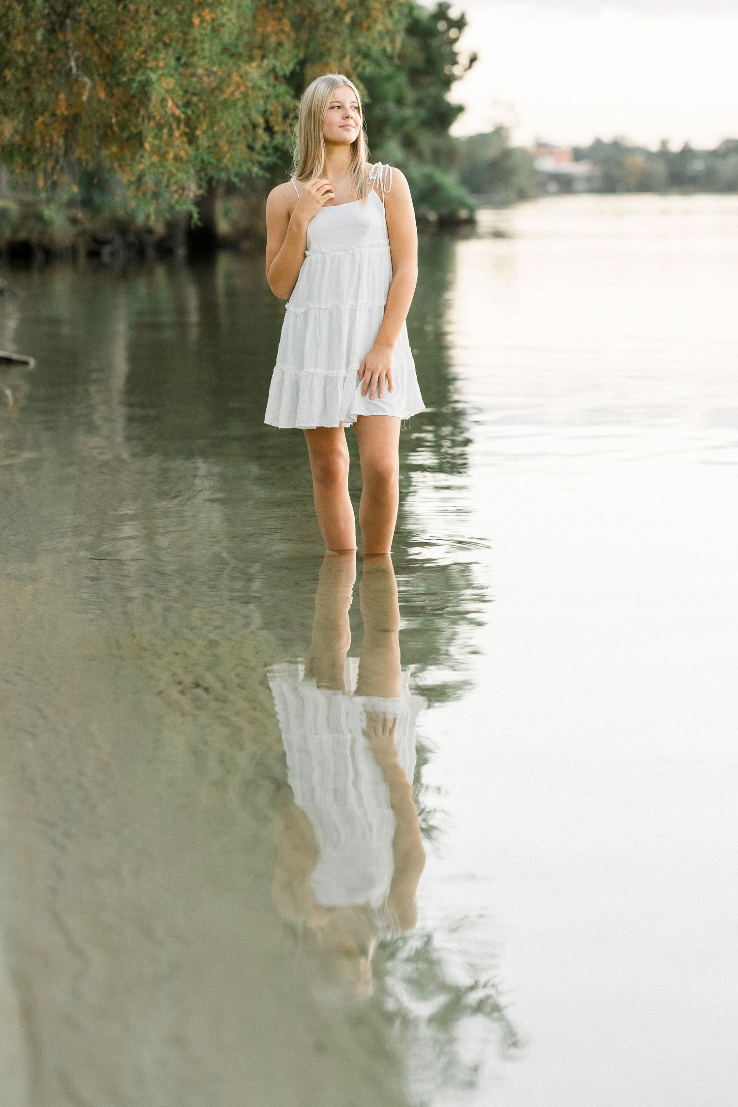 Young blonde woman in white sundress standing in shallow calm water with tree reflections, serene nature scene.
